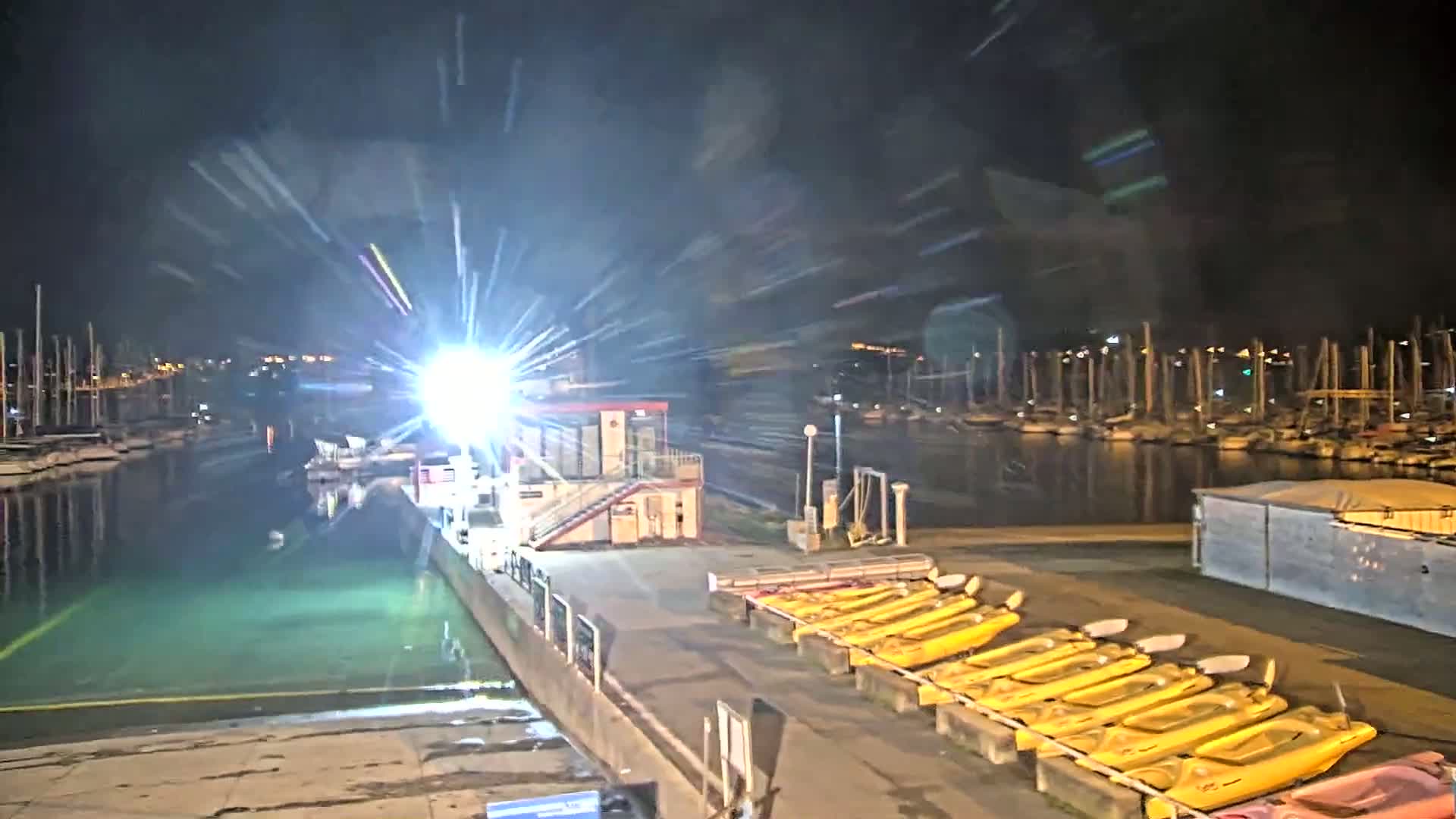 A nighttime view of a marina with numerous boats docked, a small building with lights, and a row of yellow kayaks along a dock under bright lights.