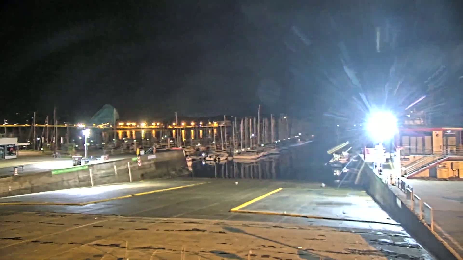 A brightly lit boat launch area at night overlooks a marina filled with docked sailboats under a dark, possibly overcast sky.