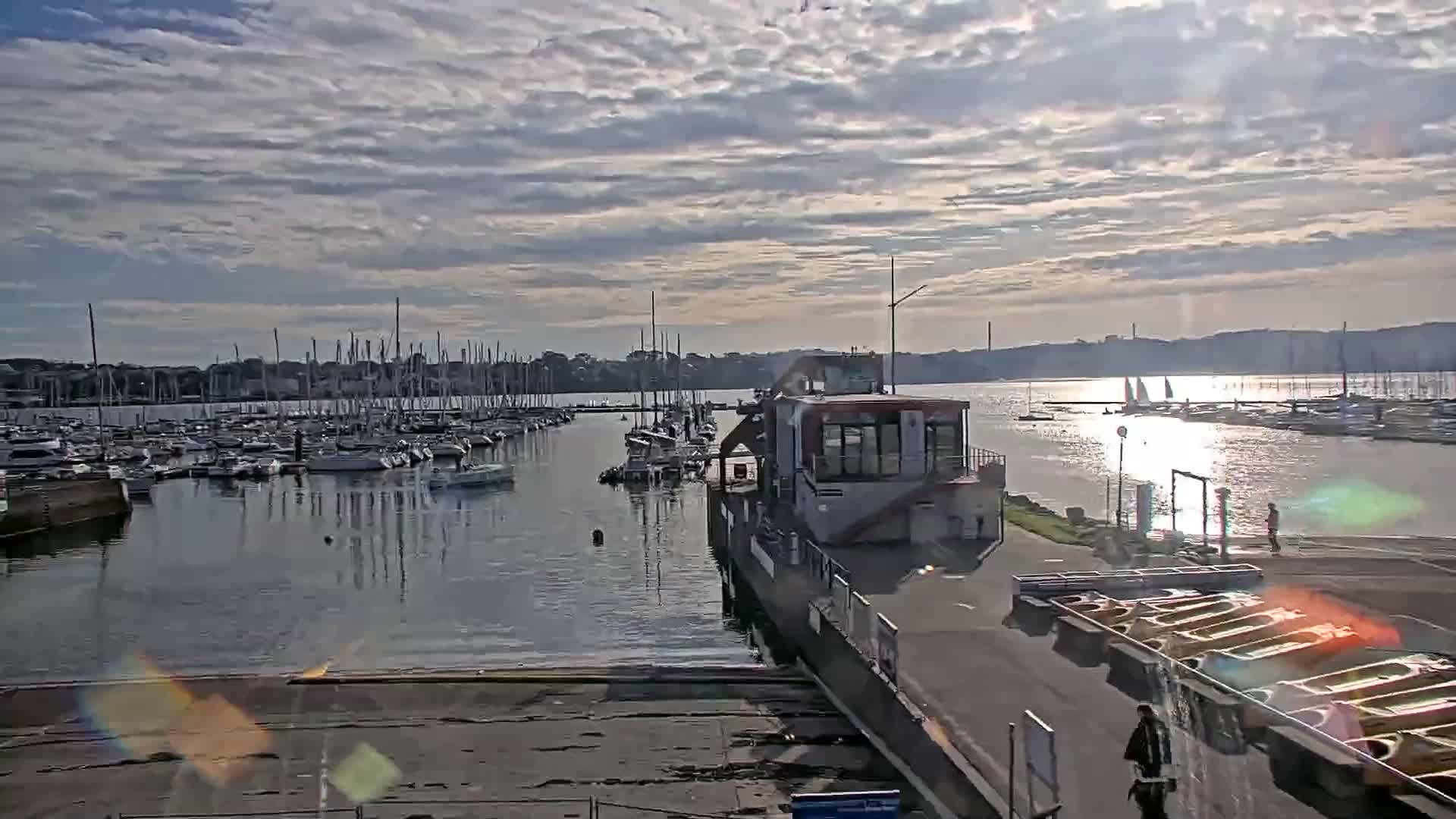 A marina filled with sailboats is seen on a partly sunny day with a cloudy sky, and a building with a boat hoist is in the foreground.