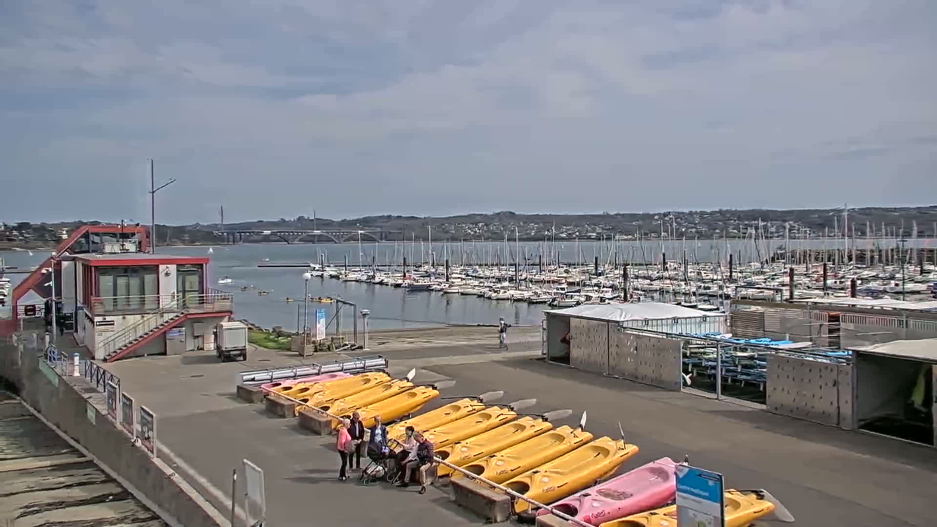 A marina with numerous sailboats and several yellow kayaks lined up on the shore, under a partly cloudy sky.