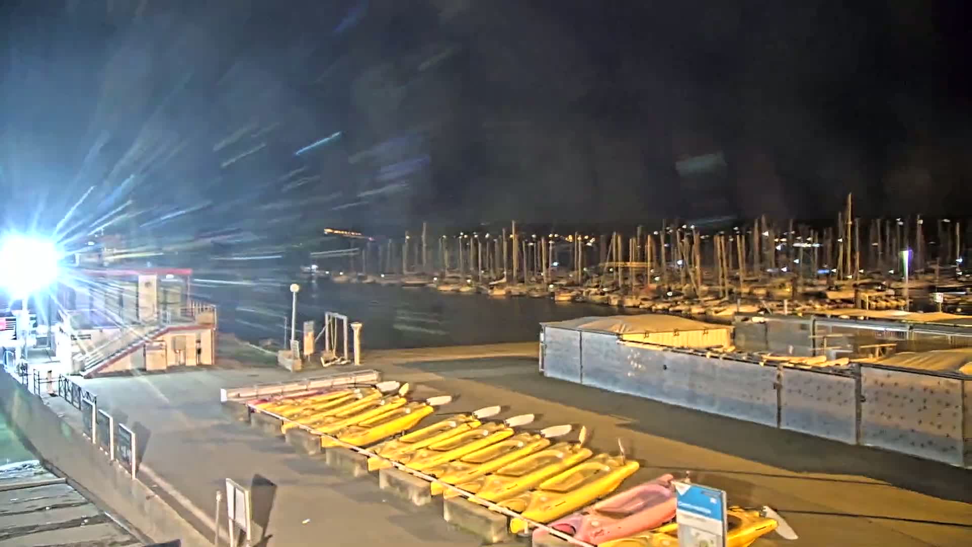 At night, a marina filled with sailboats is visible beyond a dock where numerous yellow kayaks are lined up for rental.