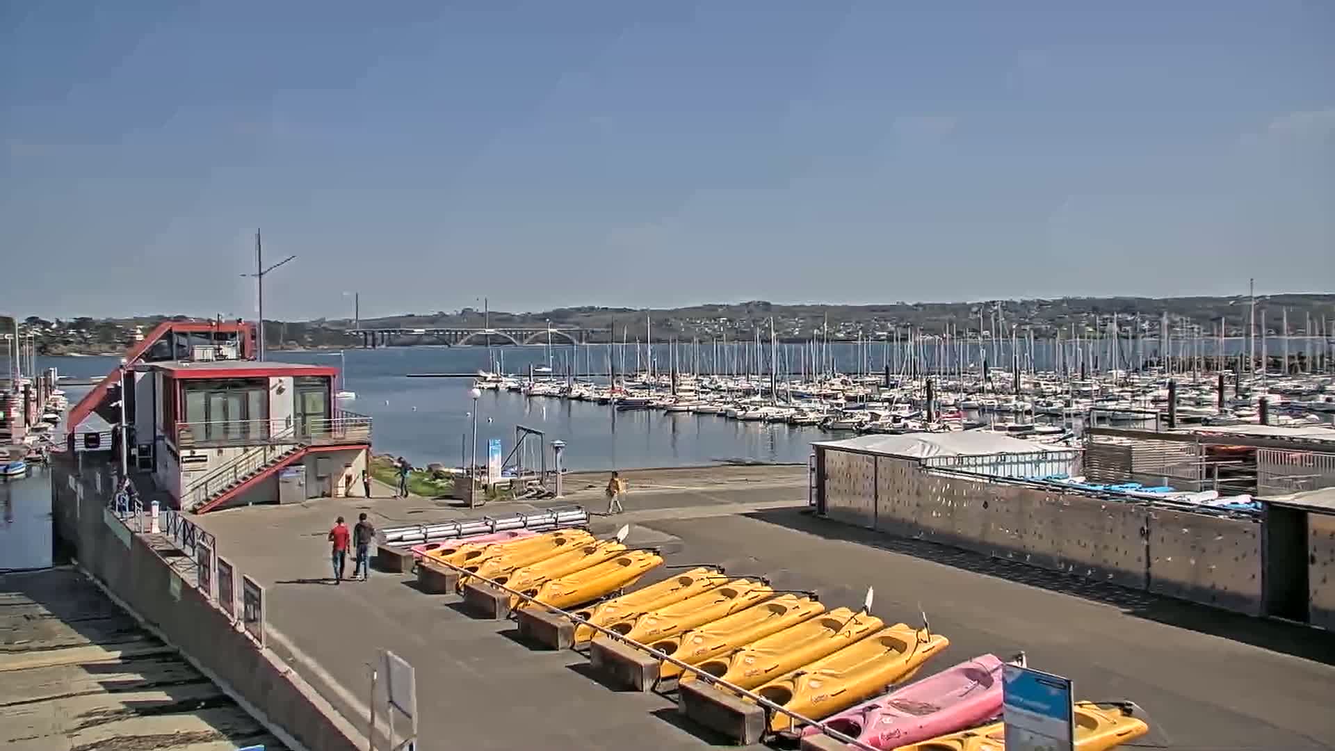 A marina with numerous sailboats and kayaks under a clear blue sky, a small building with a staircase and a few people walking nearby.