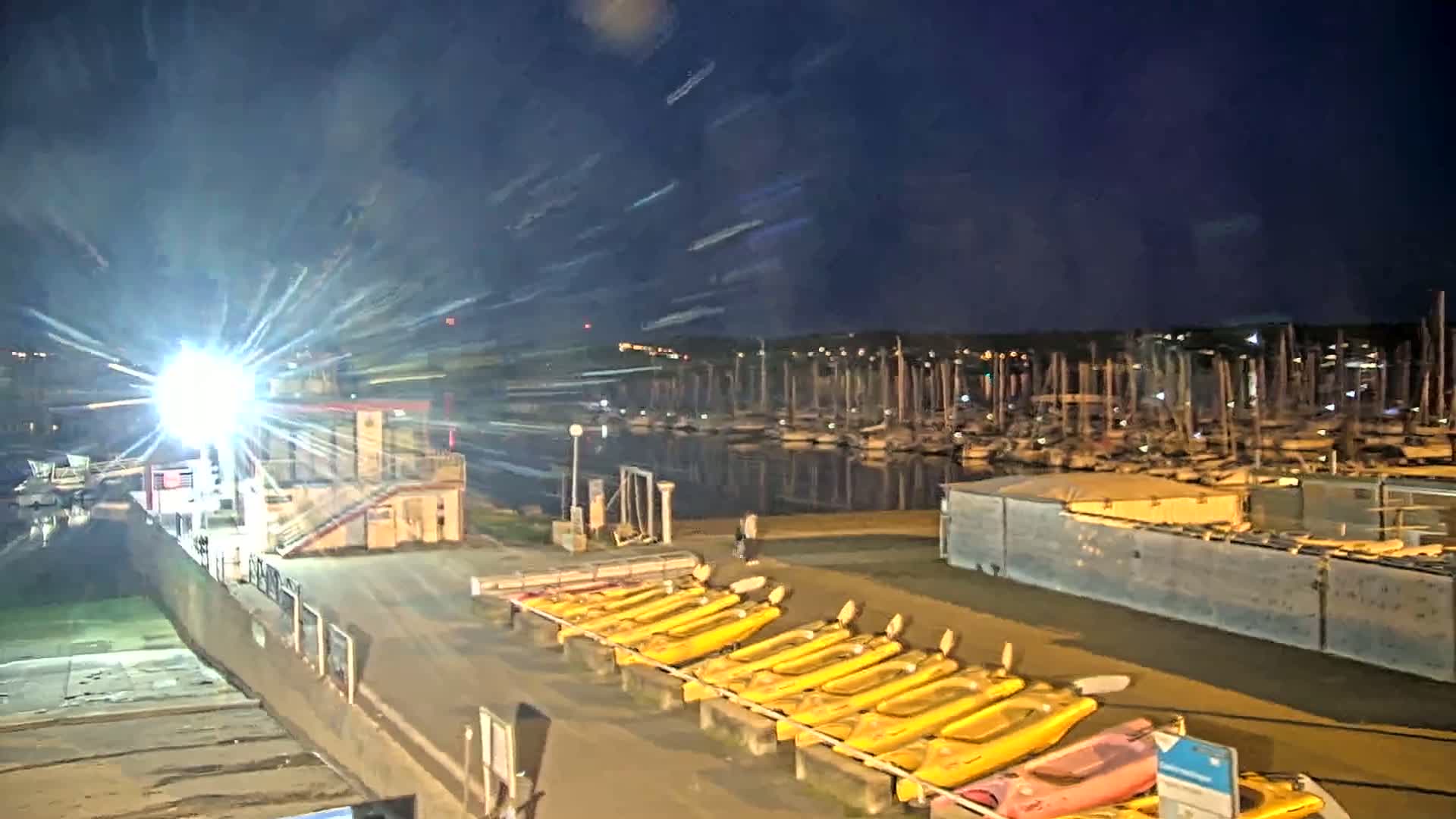 A nighttime view of a marina with numerous boats and yellow kayaks lined up on a dock under a dark, clear sky.