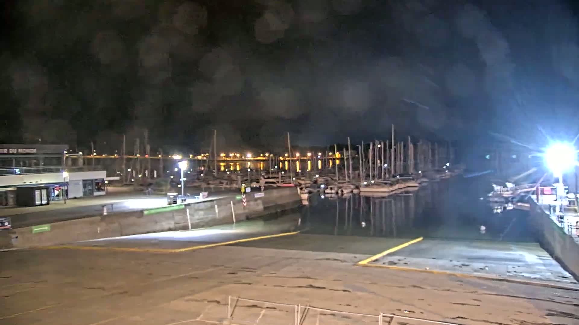 A nighttime view of a marina with numerous boats docked, illuminated by artificial lights under a dark, cloudy sky.