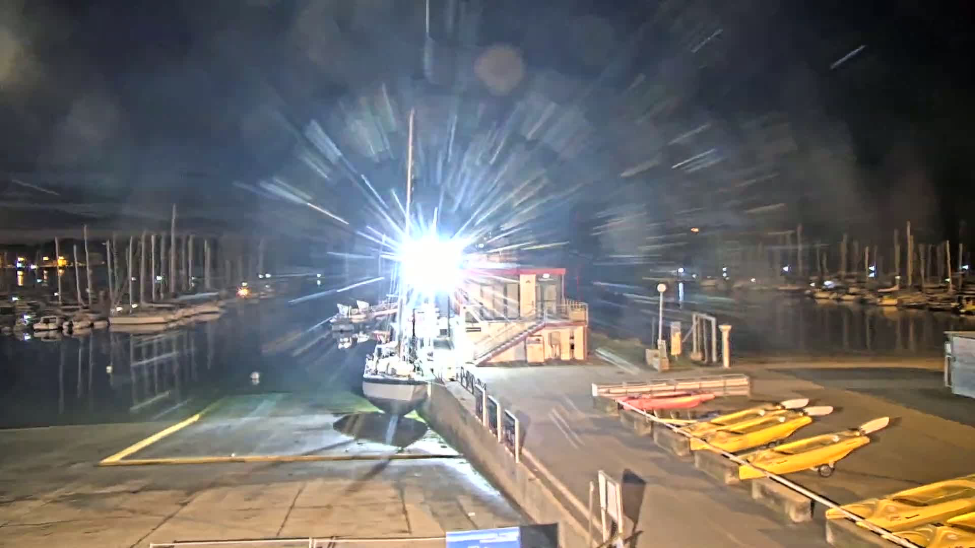A brightly lit sailboat is docked at a marina at night, with numerous other boats visible in the background and several kayaks stored nearby.