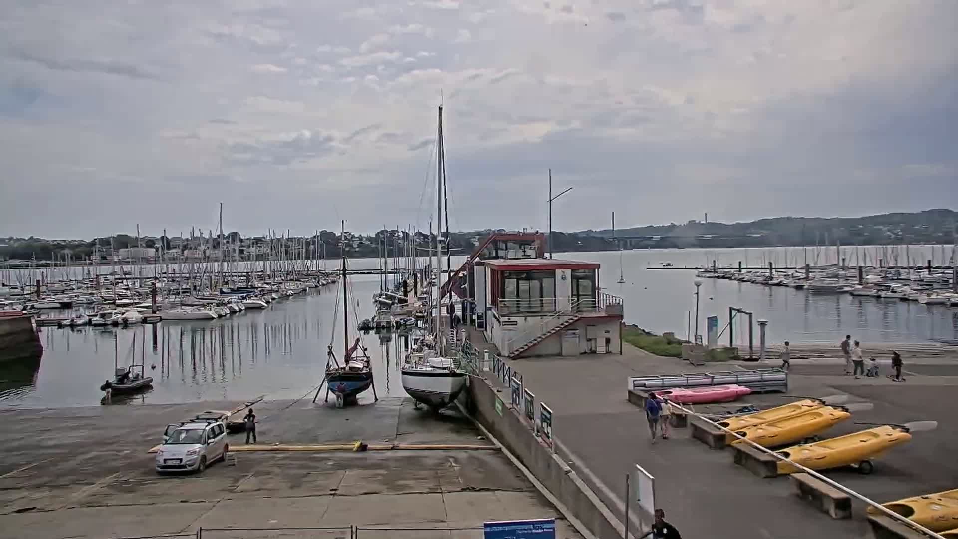 A marina filled with sailboats under a cloudy sky, with a small building and several kayaks on the shore.