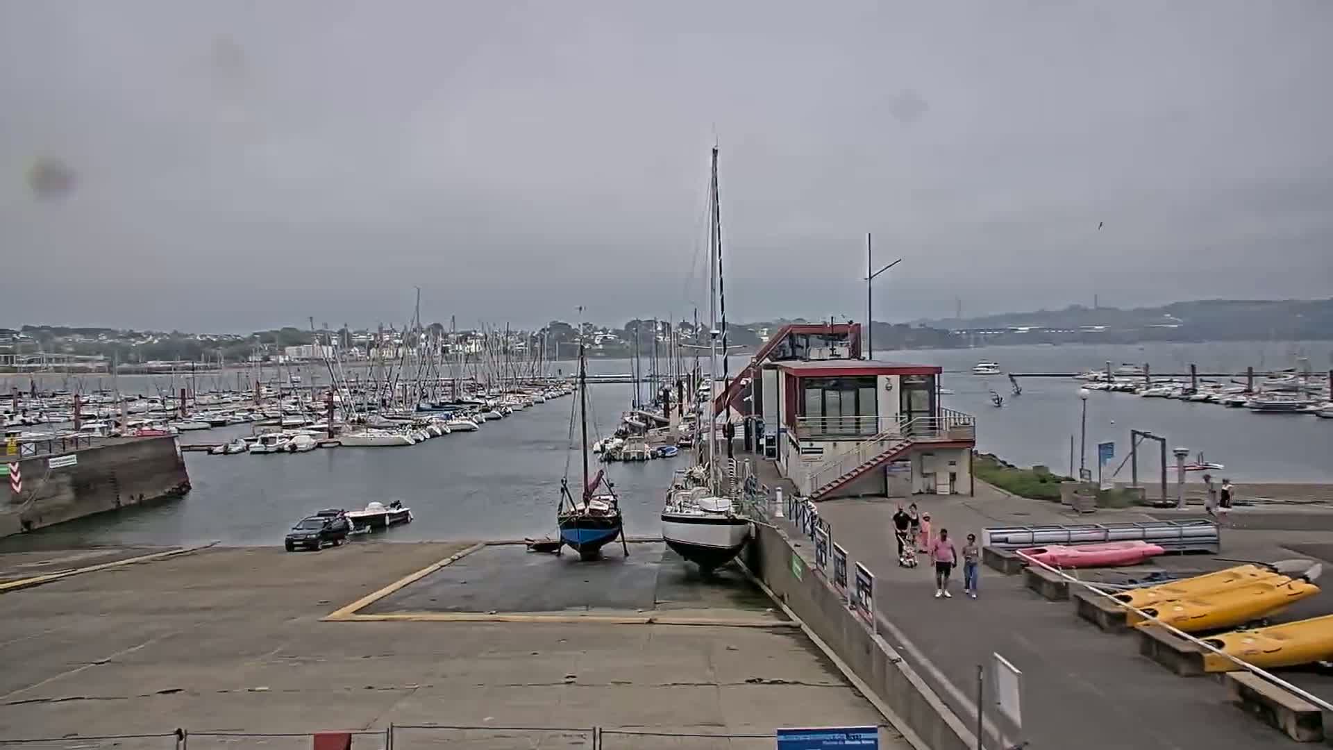 A marina with numerous sailboats and motorboats under an overcast sky, showing a boat ramp with a vehicle and several people walking near kayaks.