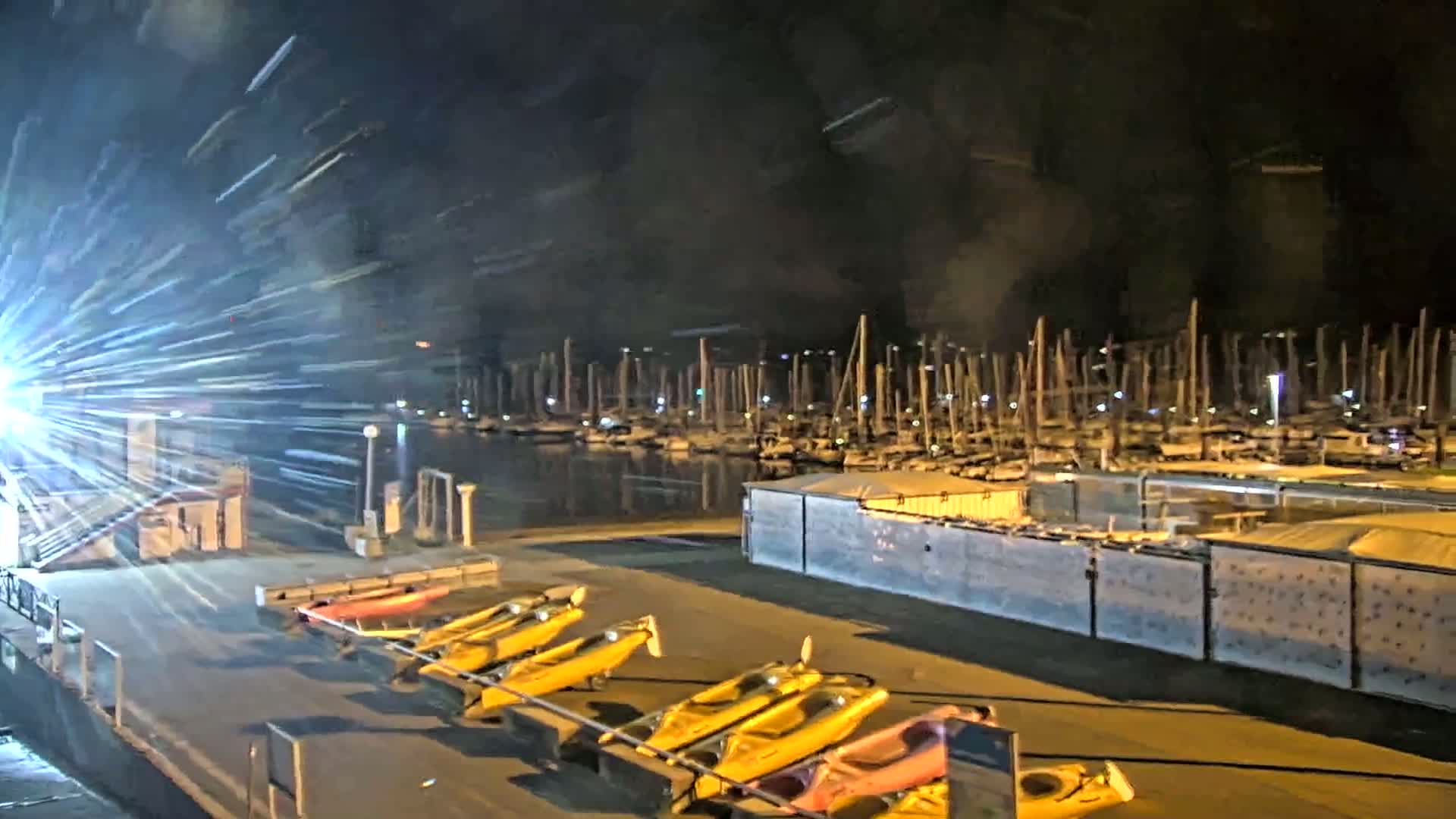 Several yellow kayaks are lined up on a dock at night next to a marina full of sailboats under a dark sky with bright light streaks.