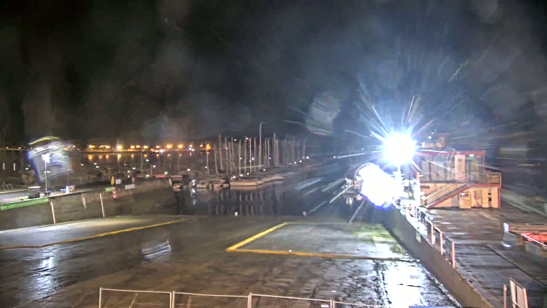 A bright light shines from a structure near a wet dock at night, illuminating moored boats and a waterfront area.