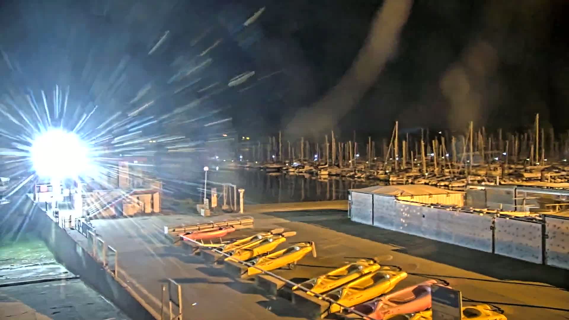 At night, a bright light shines on a dock with yellow kayaks lined up near a marina filled with sailboats under a dark, possibly smoky or hazy sky.