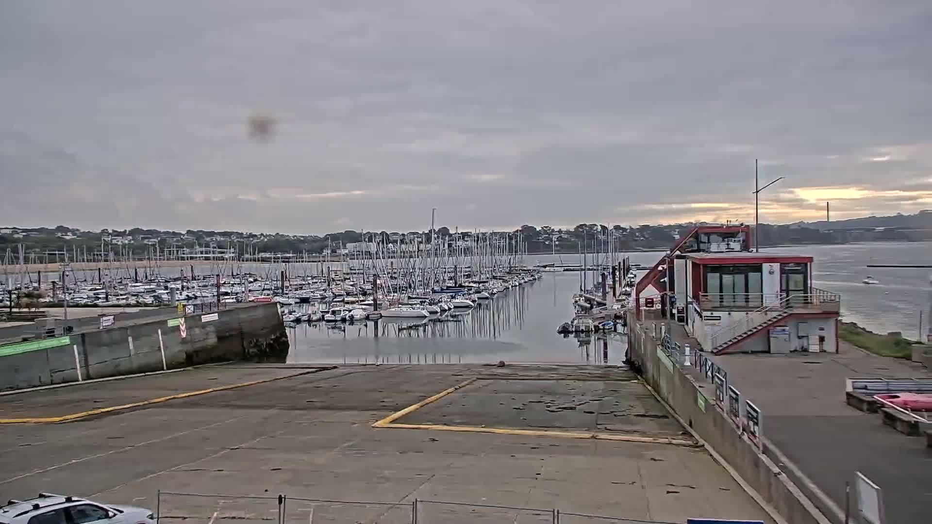 A marina filled with sailboats is viewed from a paved area leading to the water under an overcast sky.