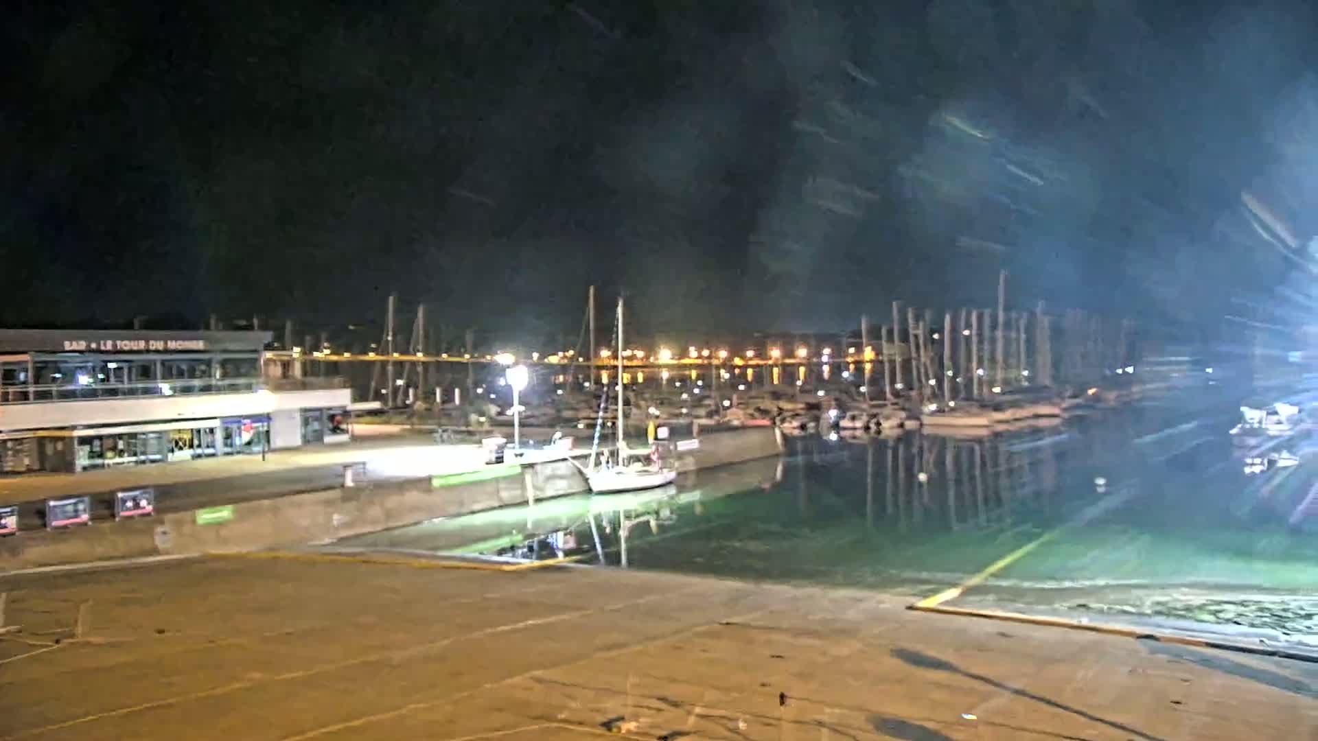 A nighttime view of a marina with numerous boats docked, a building with lights on, and green-tinged water.