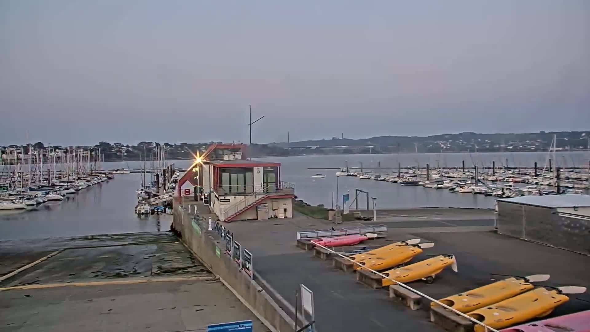 A marina filled with sailboats is seen under a dusky sky, with several kayaks lined up on the shore near a small building.