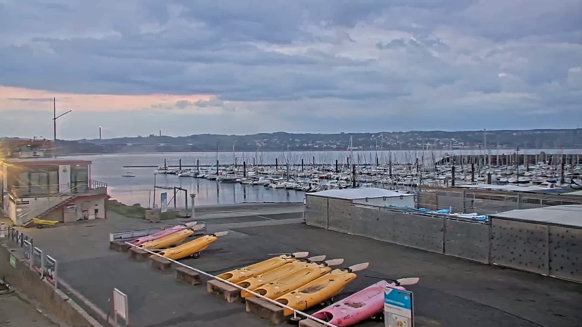 Under a cloudy sky, a marina filled with sailboats is visible across calm water from a waterfront area where several yellow and pink kayaks are stored.