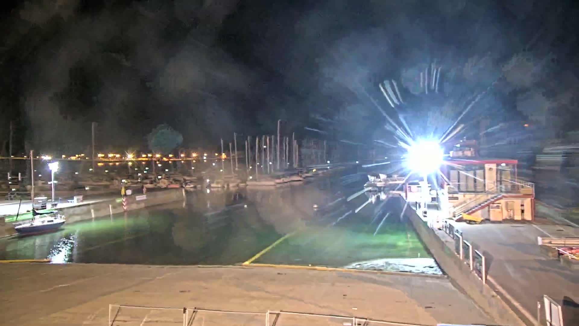 A brightly lit marina at night, with several boats docked, is partially obscured by bright light flares and hazy atmospheric conditions.