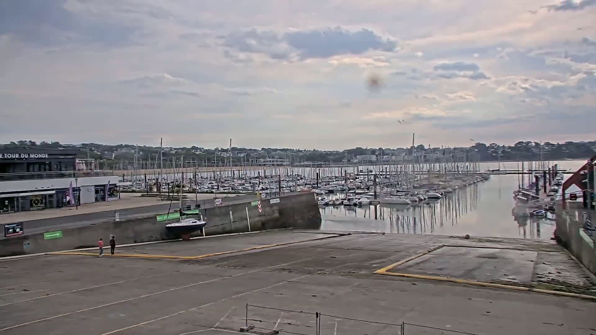 A marina filled with sailboats under a cloudy sky is seen from a paved area with a single sailboat docked nearby.