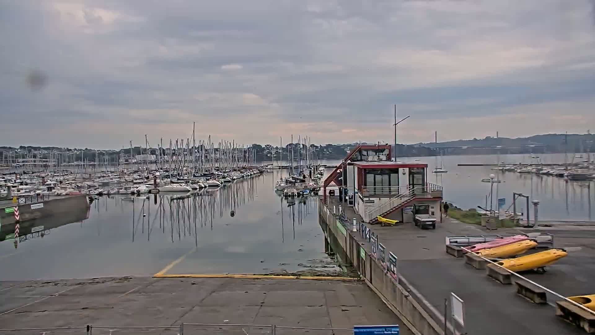 A marina filled with sailboats under a cloudy sky, with a small building and kayaks nearby.