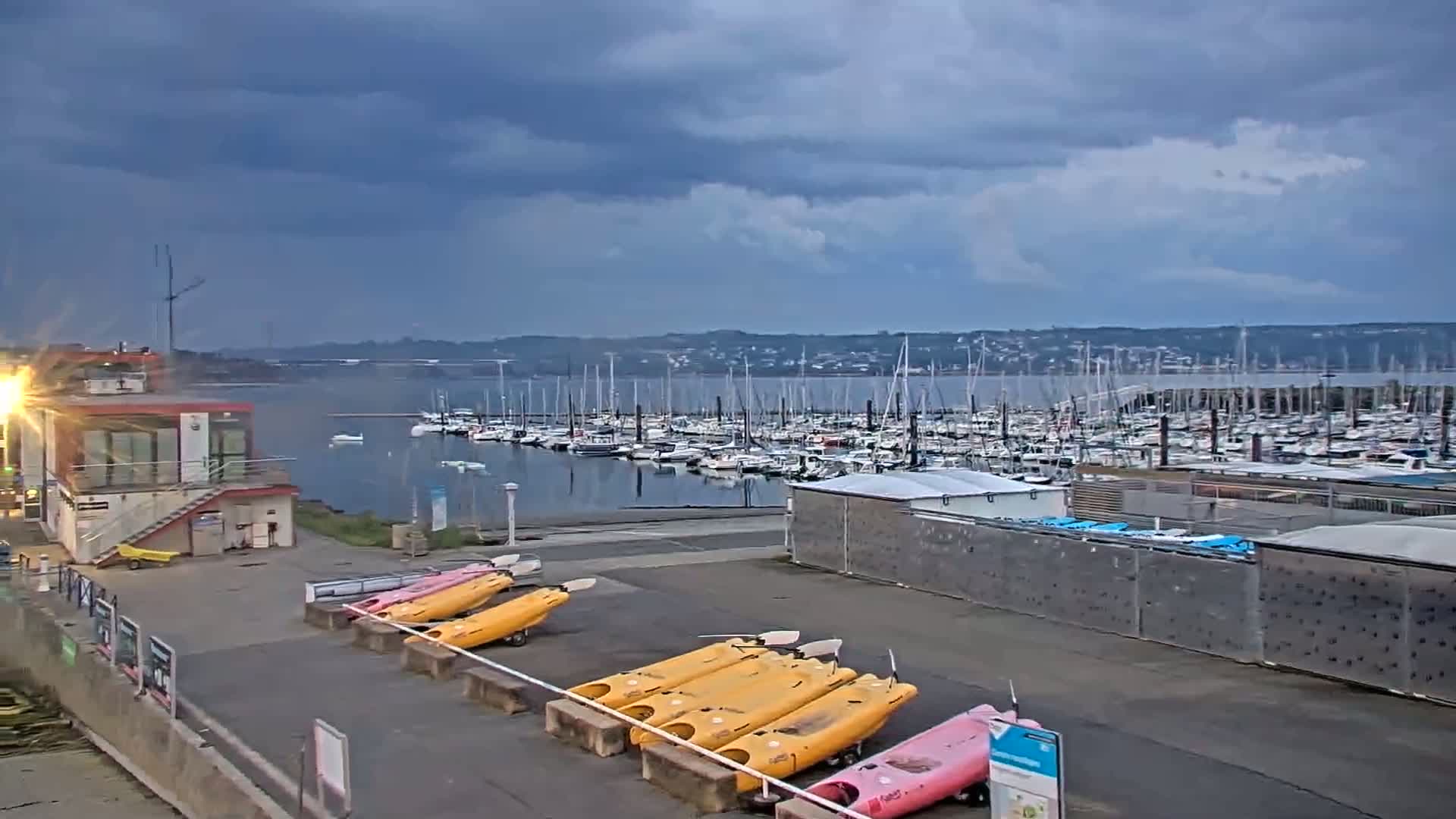 Kayaks are lined up near a small building overlooking a large marina filled with sailboats under a cloudy sky.