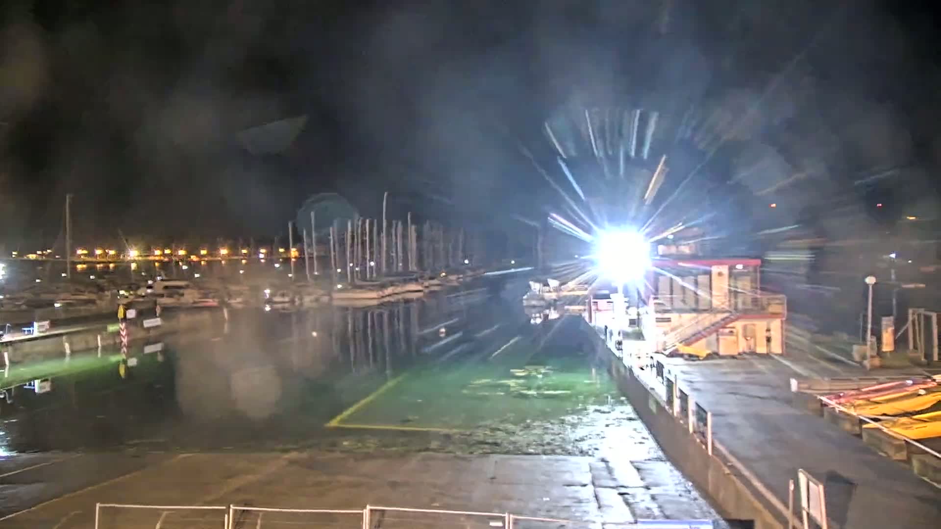A nighttime view of a marina with many boats docked, illuminated by lights, with a bright light source near a small building, and the water appearing greenish.