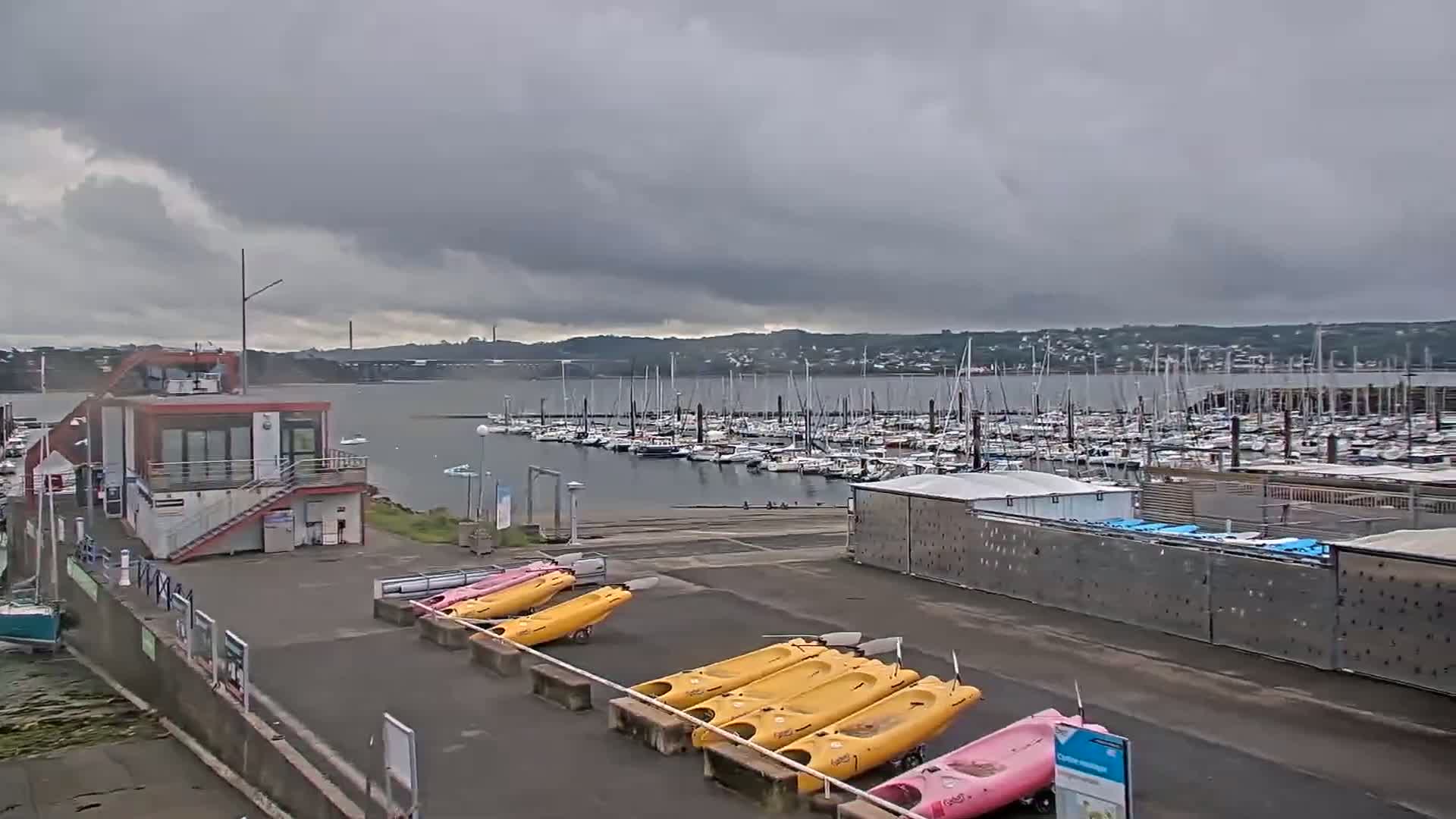 A marina with numerous boats, kayaks, and a small building is shown under an overcast sky.