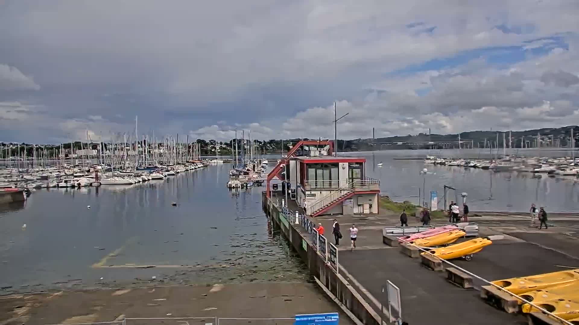 A marina filled with sailboats under a partly cloudy sky, with a small building and several people near kayaks on a nearby dock.