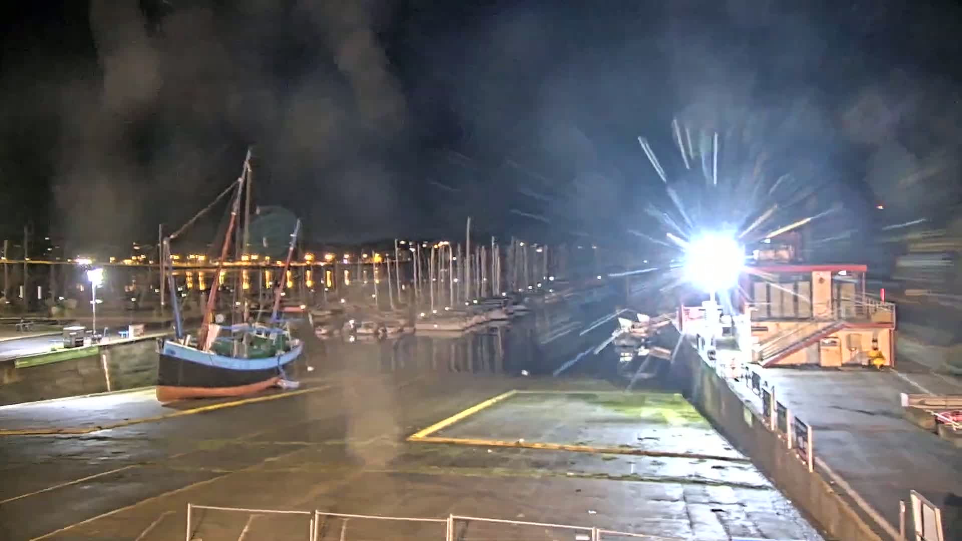 A small sailboat is docked in a marina at night, with bright lights and smoke or fog in the air.