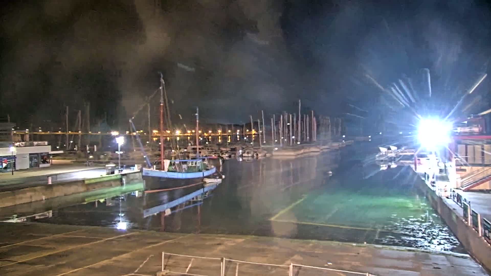 A nighttime view of a harbor with a docked sailboat and many masts in the background, under a dark, smoky sky, showing bright artificial light illuminating the water.