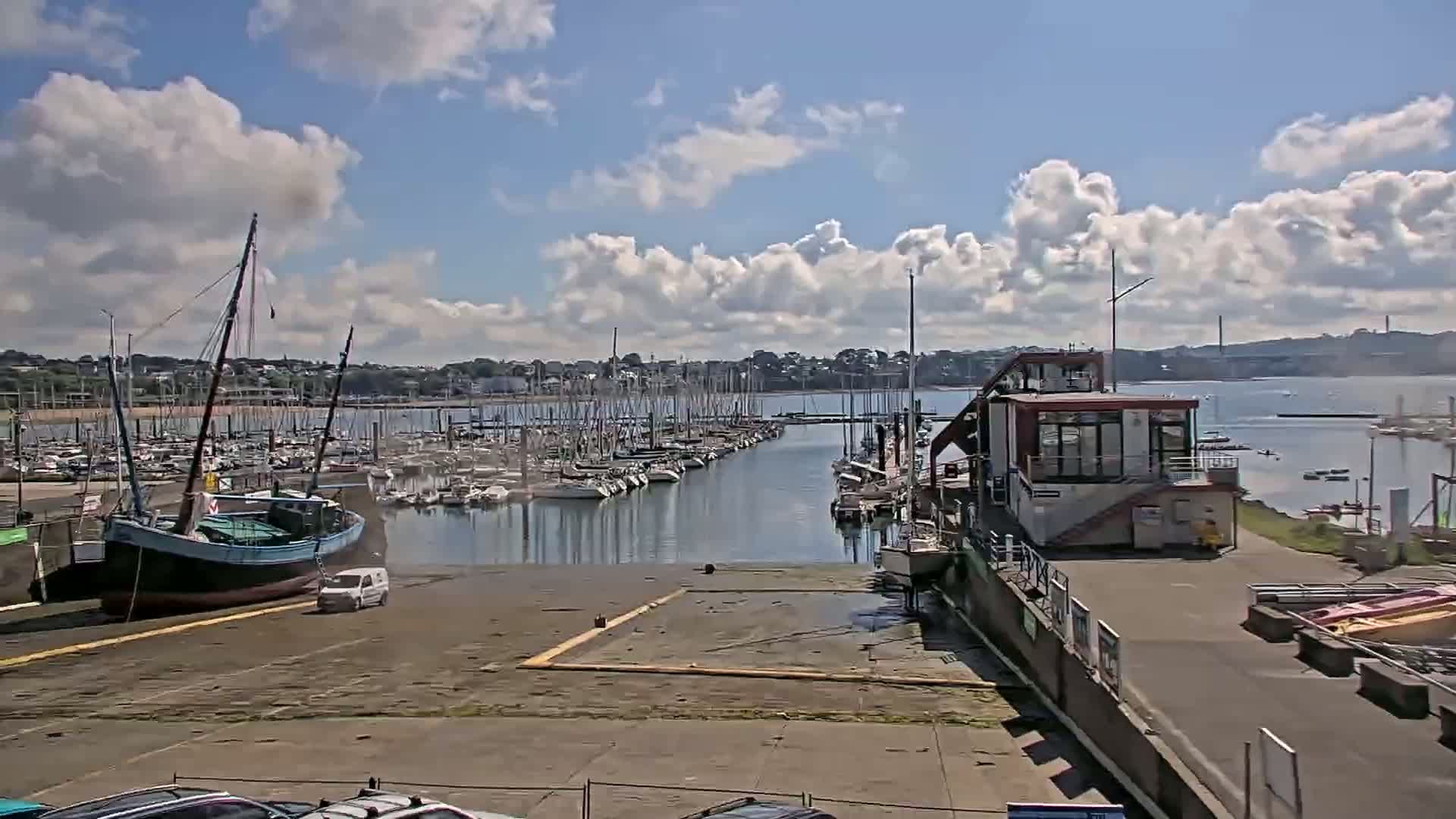 A marina filled with sailboats under a partly cloudy sky, with a large wooden ship and a small van parked near the docks.