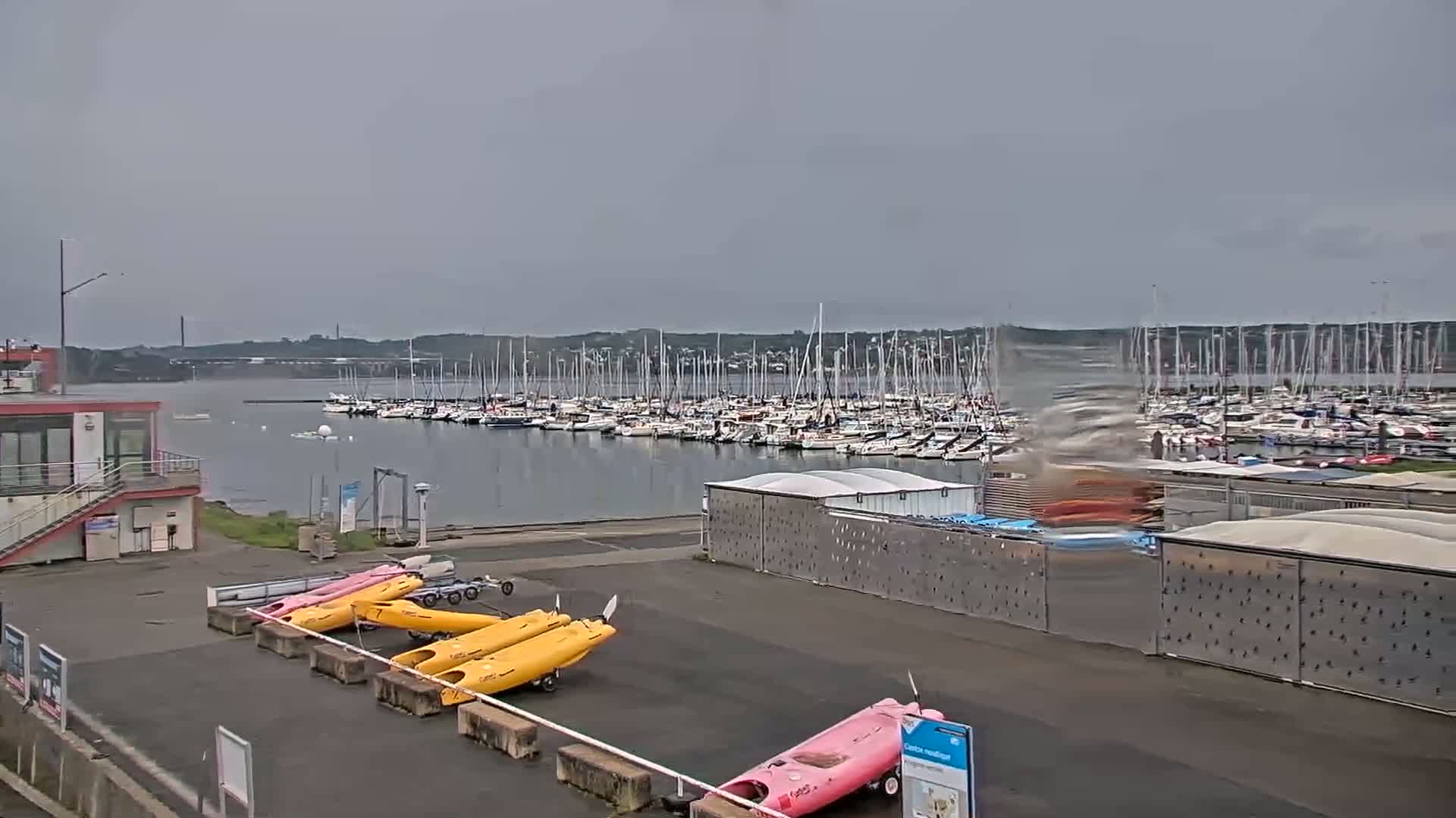 A large marina filled with numerous sailboats is visible across a calm body of water under an overcast sky, with kayaks and other small watercraft stored nearby on land.