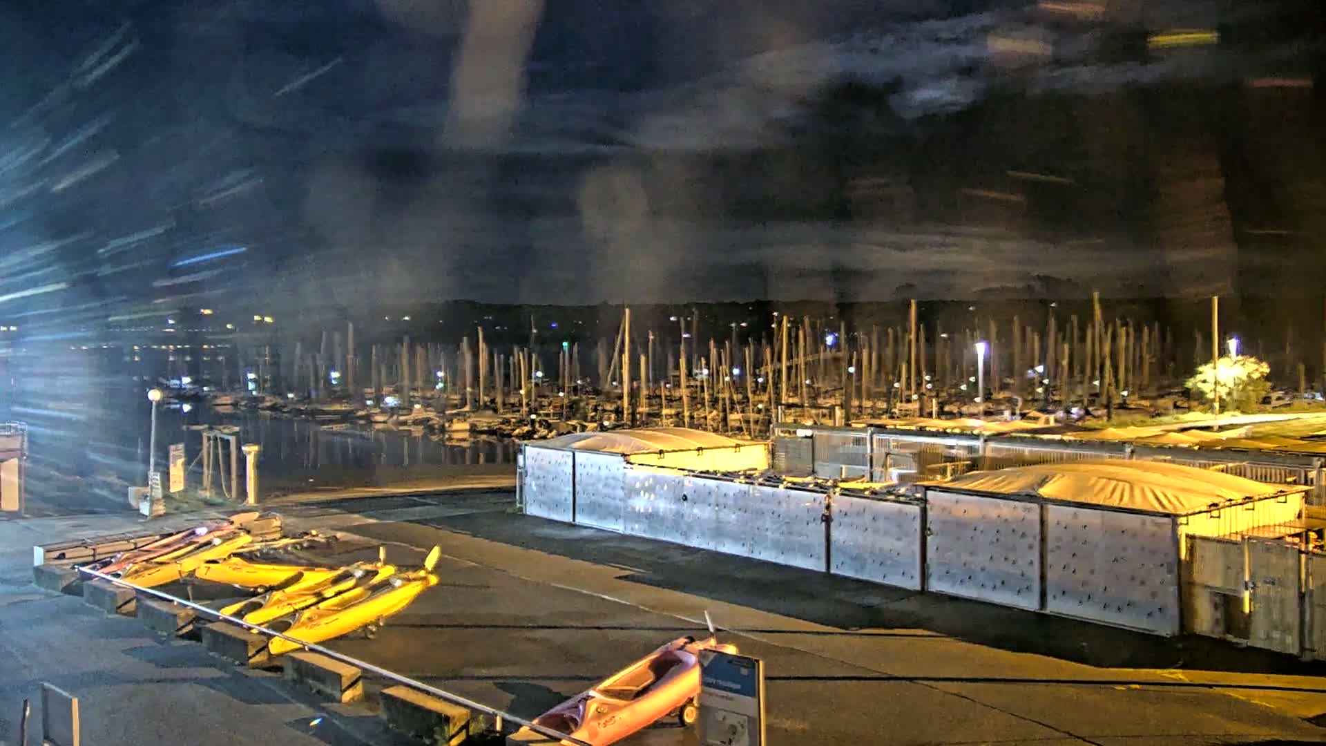 At night, under a cloudy sky, a marina filled with numerous boats is visible beyond a fenced area containing several covered storage containers, and a few kayaks are parked nearby.