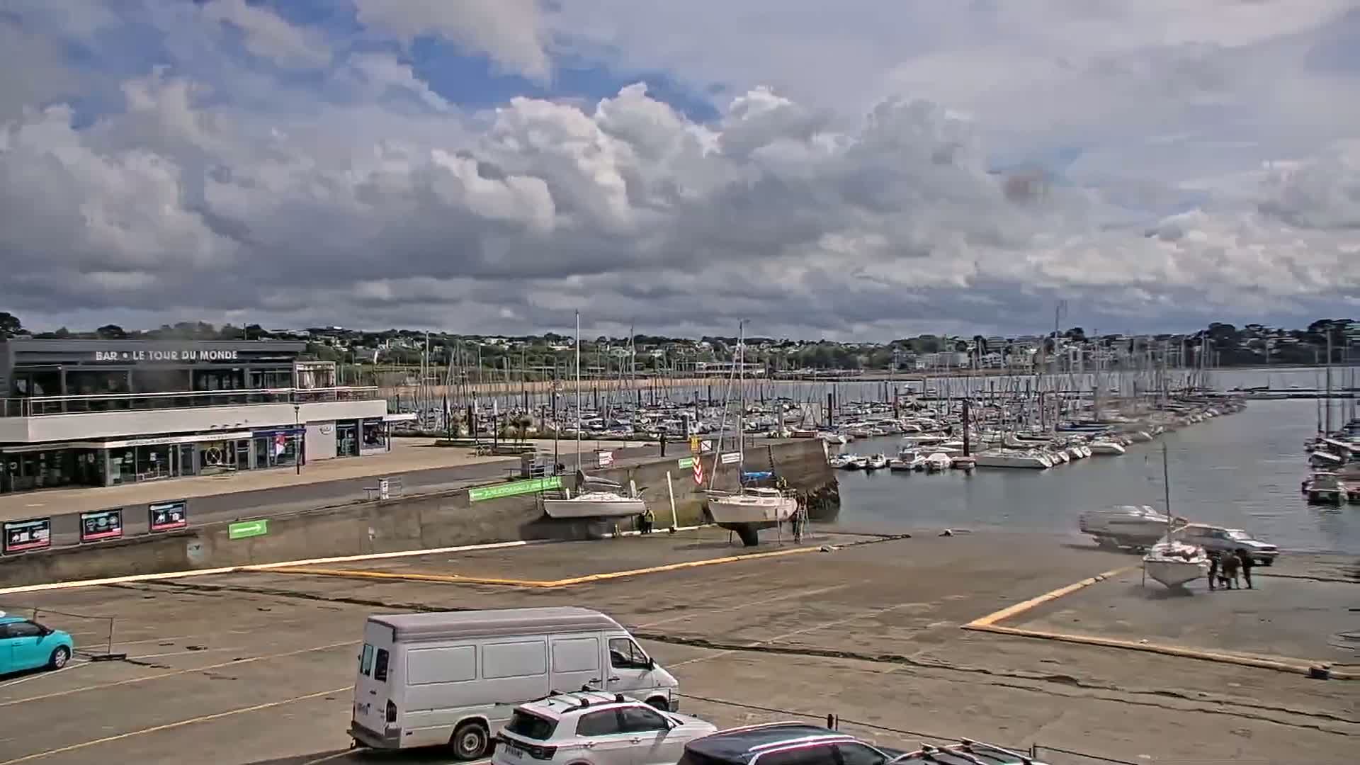 A partly cloudy day reveals a marina filled with sailboats and yachts, a building with shops, and a parking lot with several vehicles.
