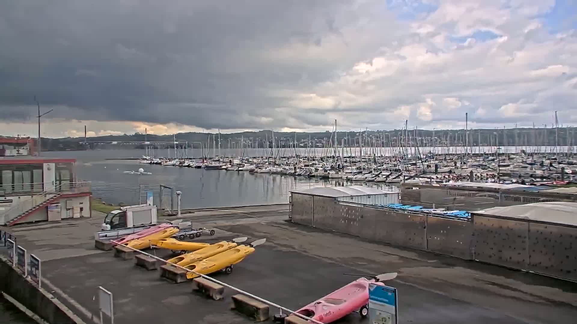 A marina filled with numerous sailboats under a cloudy sky, with several kayaks and a small utility vehicle parked nearby.