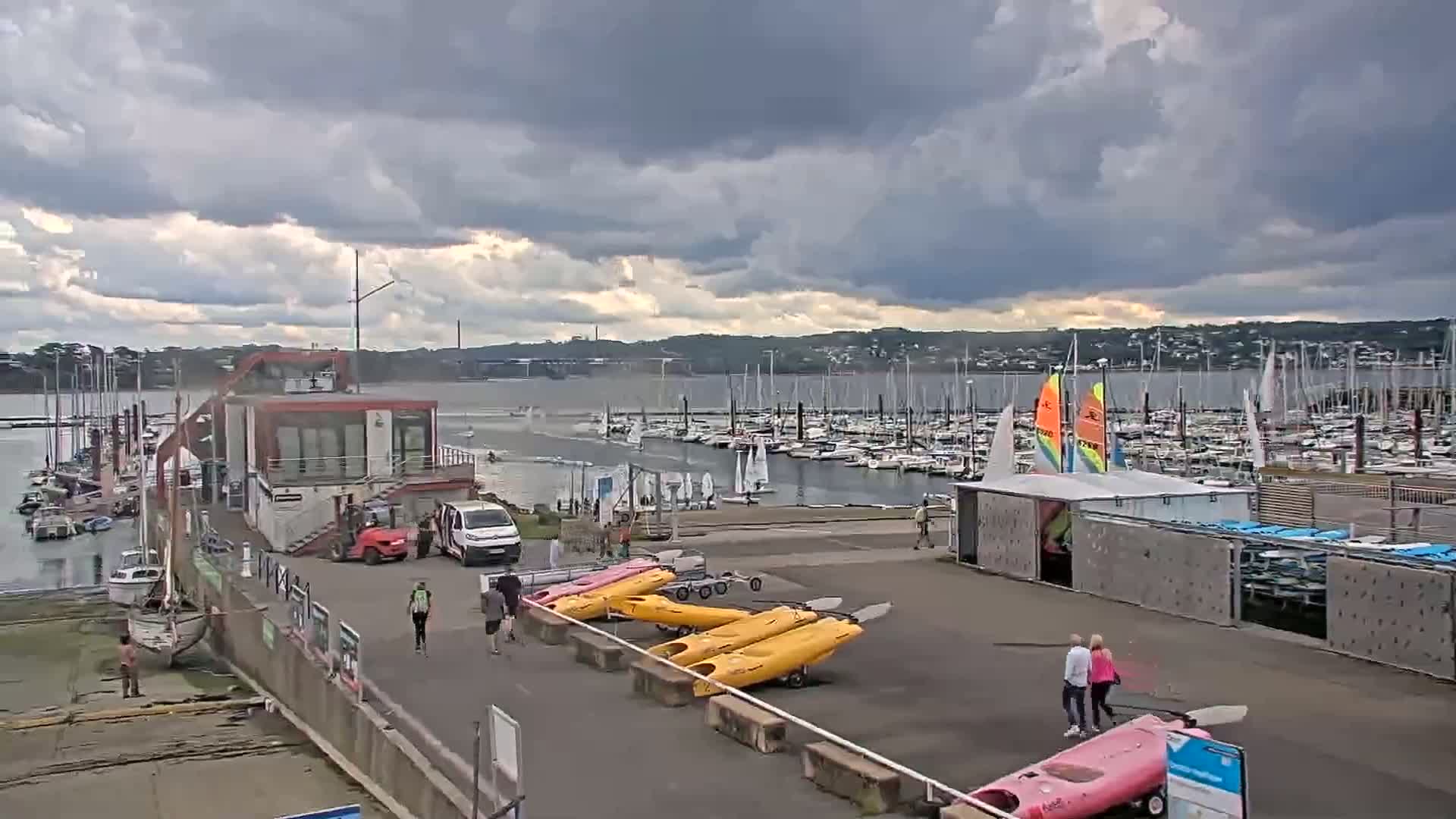 A marina with numerous sailboats and kayaks, a small building, and several people under a cloudy sky.
