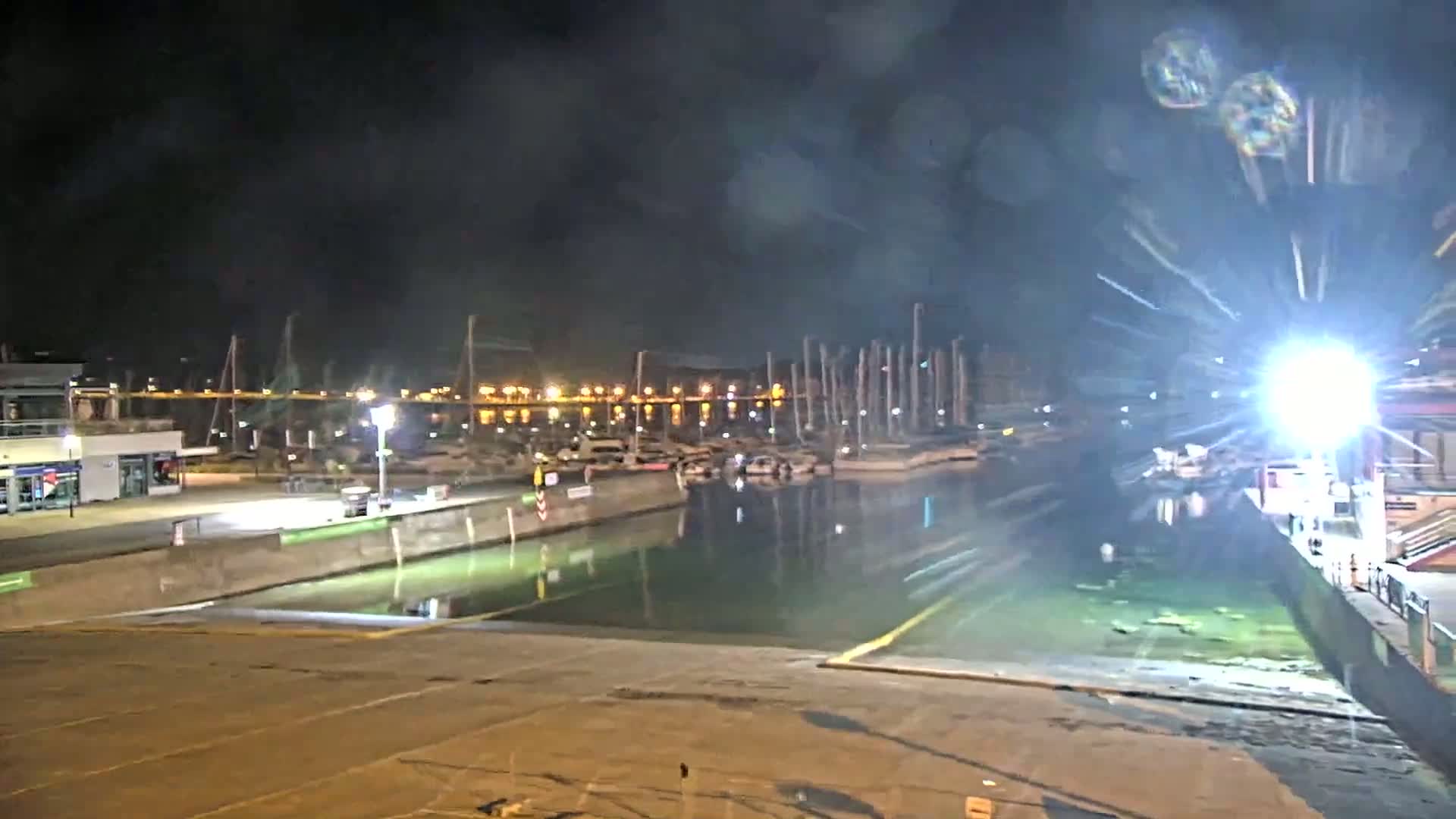 A nighttime view of a marina with docked sailboats and a lock, illuminated by bright lights, under a dark, possibly hazy sky.