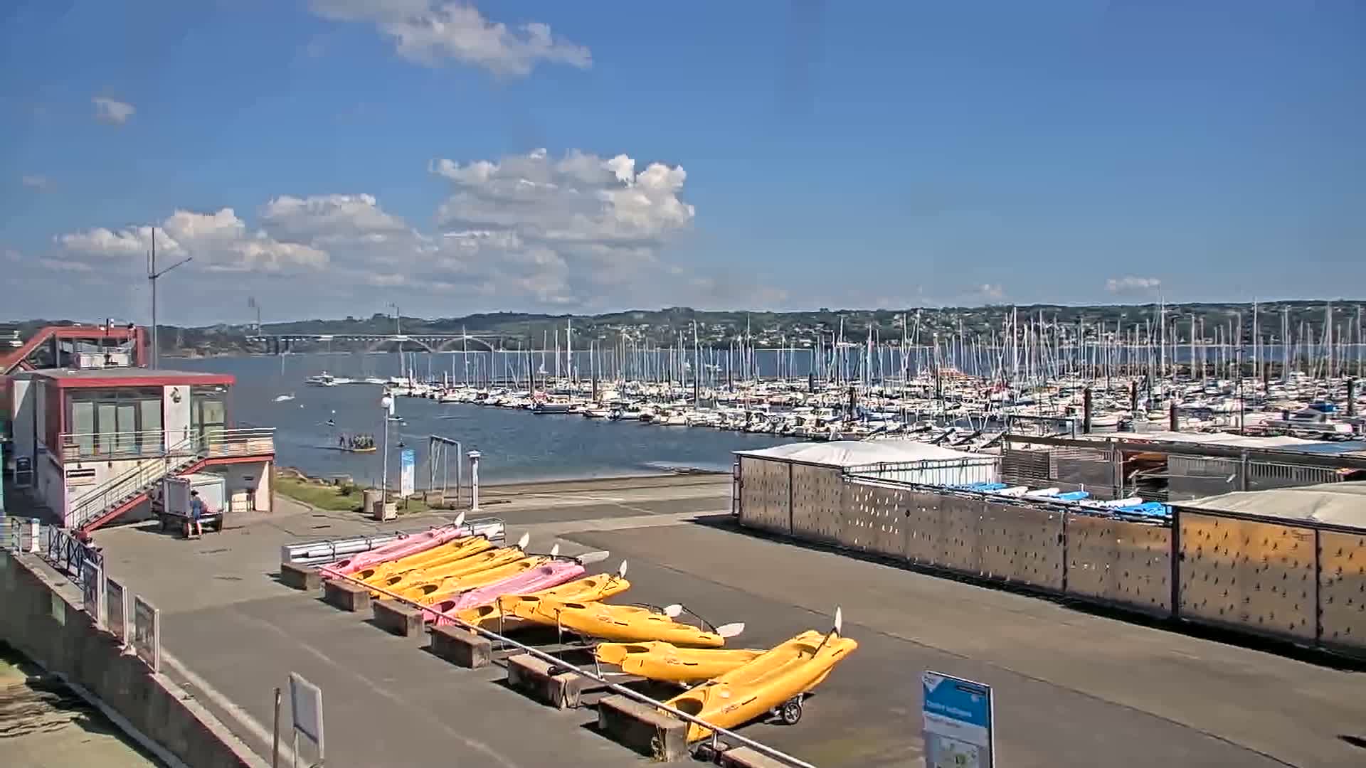 A marina with numerous boats, a small building, and kayaks is shown under a partly cloudy sky.