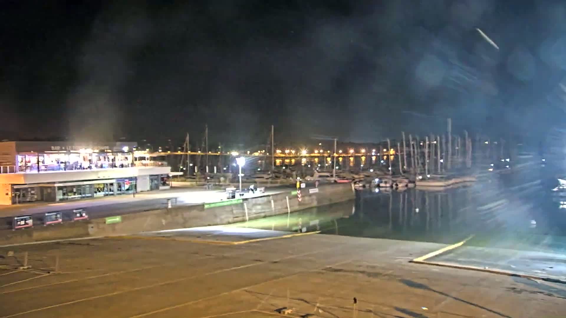 A nighttime view of a marina with a building containing a restaurant, boats moored at the docks, and hazy smoke or fog in the air.