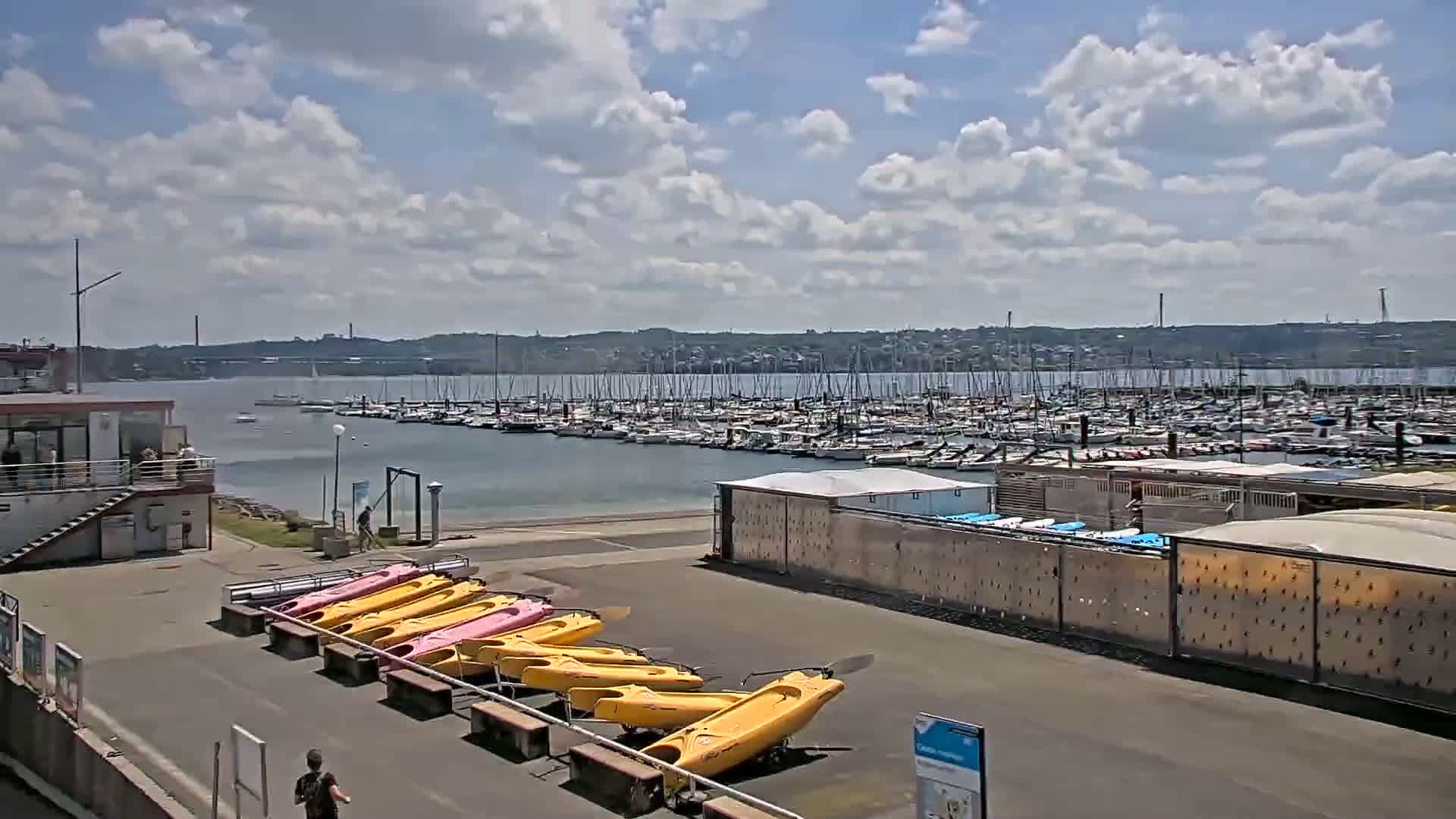 A marina with numerous sailboats and motorboats under a partly cloudy sky, with a row of kayaks in the foreground.