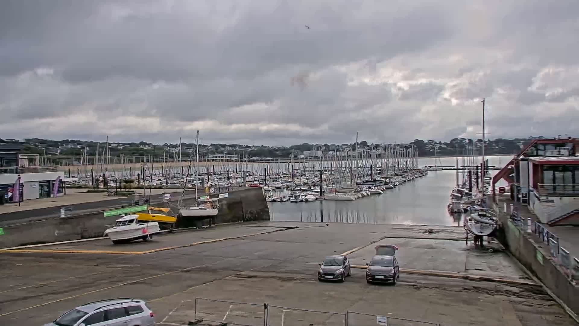 A marina filled with numerous sailboats and motorboats under a cloudy sky, with several cars parked in a nearby lot.
