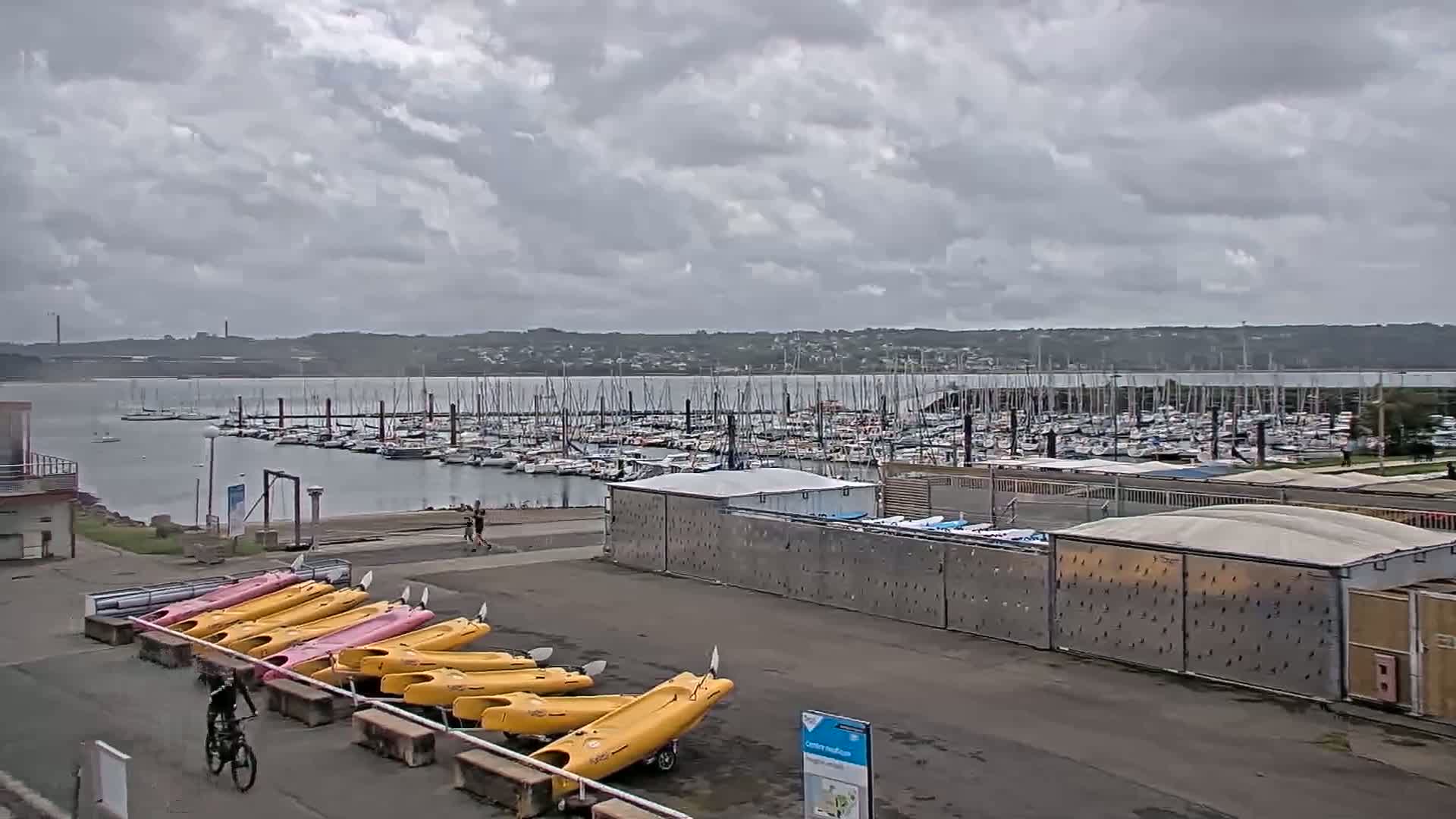 A large marina filled with sailboats is visible under a cloudy sky, with kayaks and a bicyclist in the foreground.