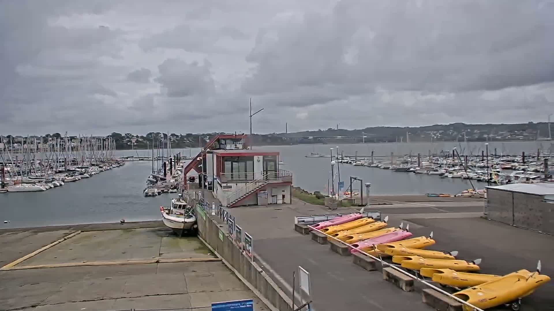A marina with numerous sailboats and motorboats moored under a cloudy sky, featuring a small building and rows of kayaks on the shore.