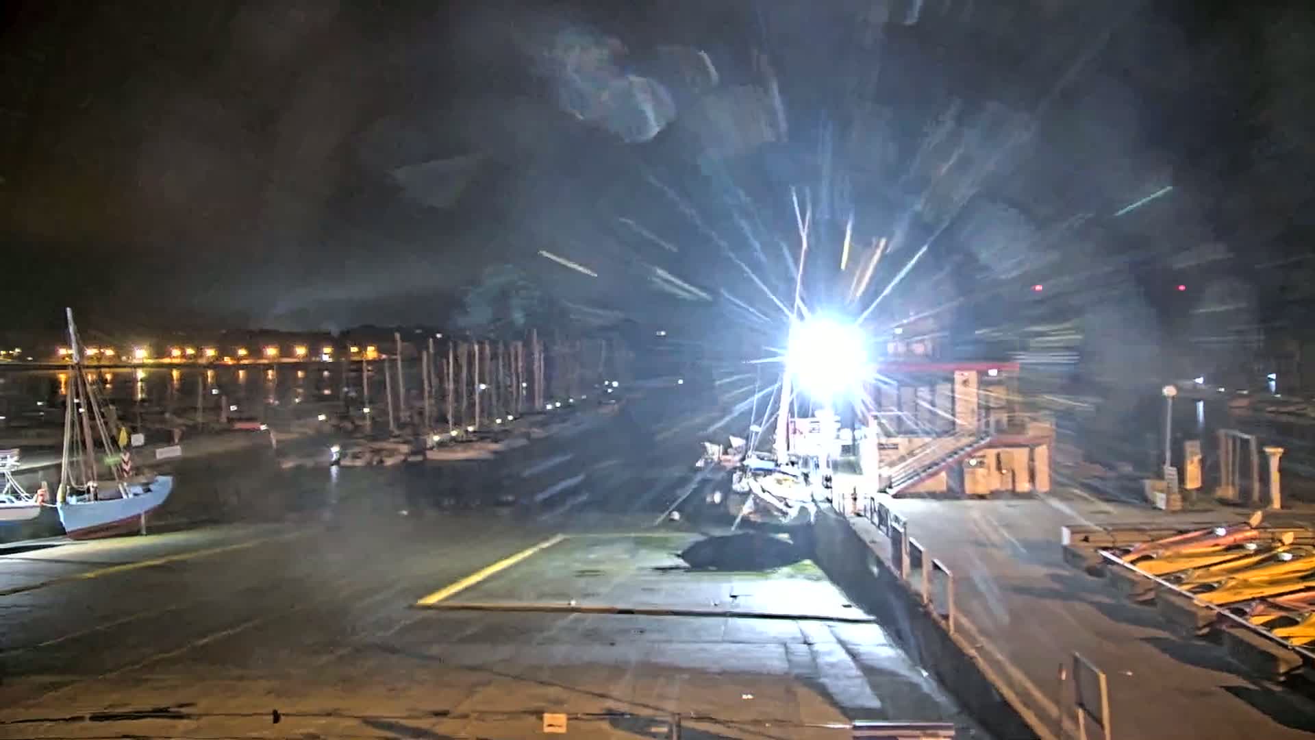 A brightly lit marina at night, with numerous boats docked, and a strong light source causing lens flare.