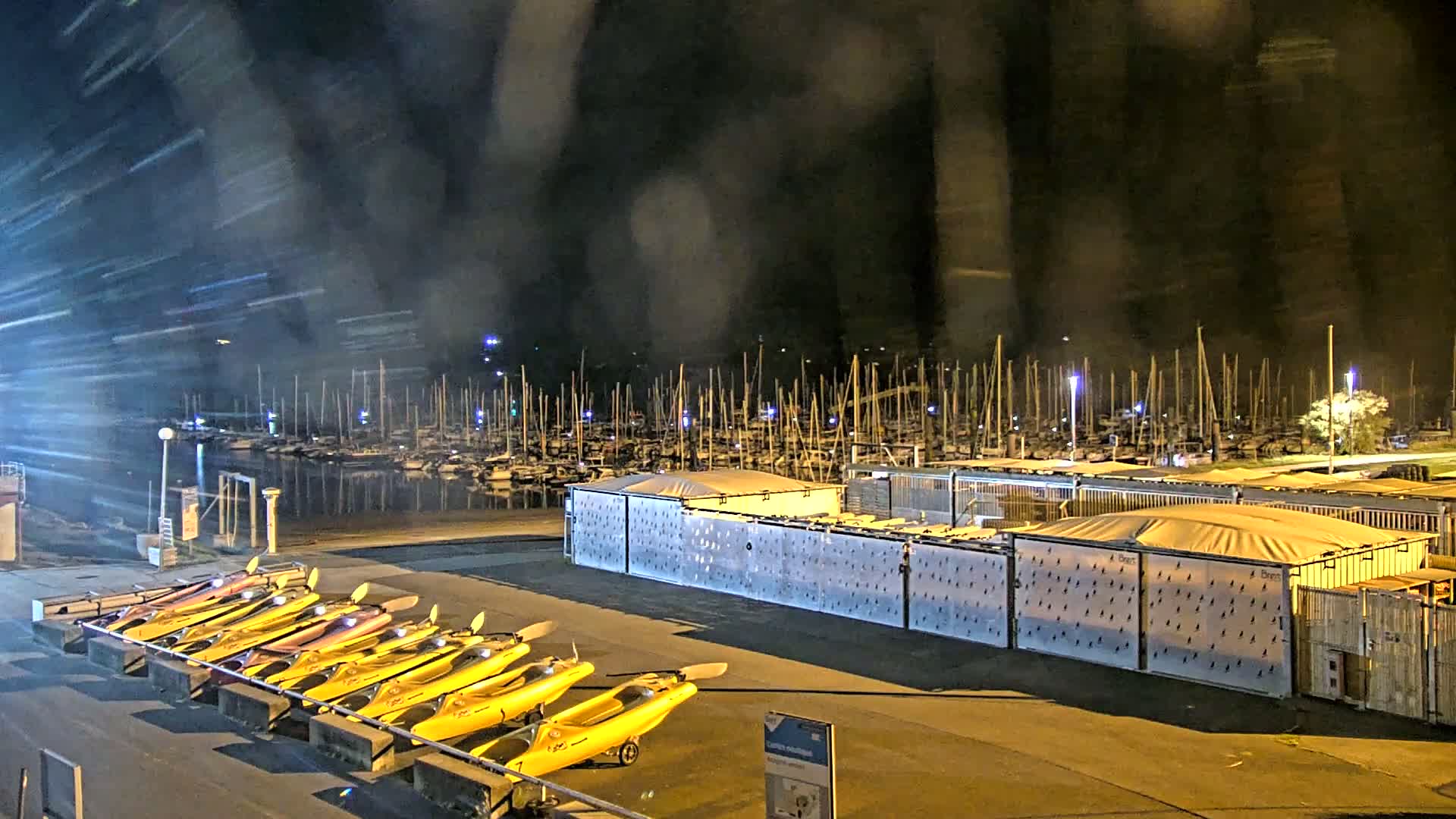 A nighttime view of a marina with many boats docked, a row of yellow kayaks, and storage containers, all under a rainy sky.
