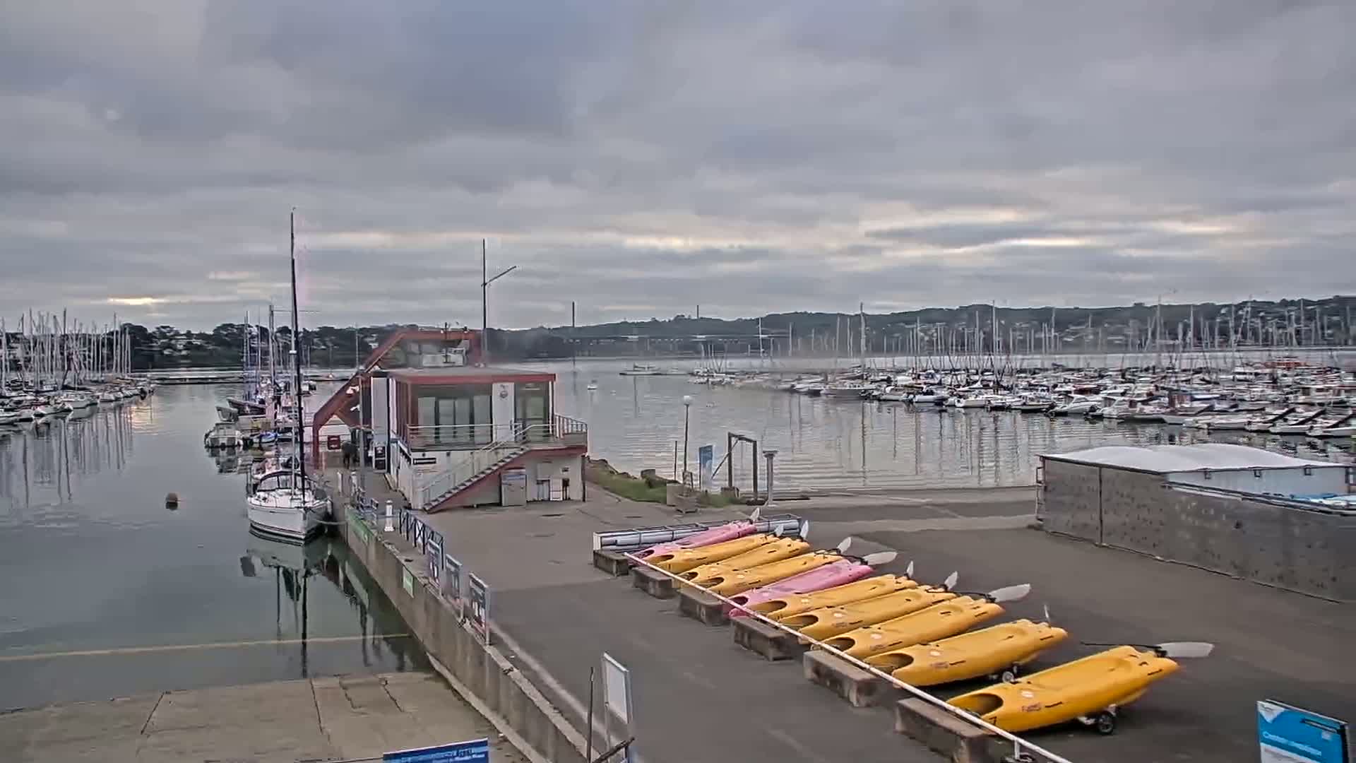 A marina filled with sailboats under a cloudy sky, with a row of kayaks lined up on the shore next to a small building.