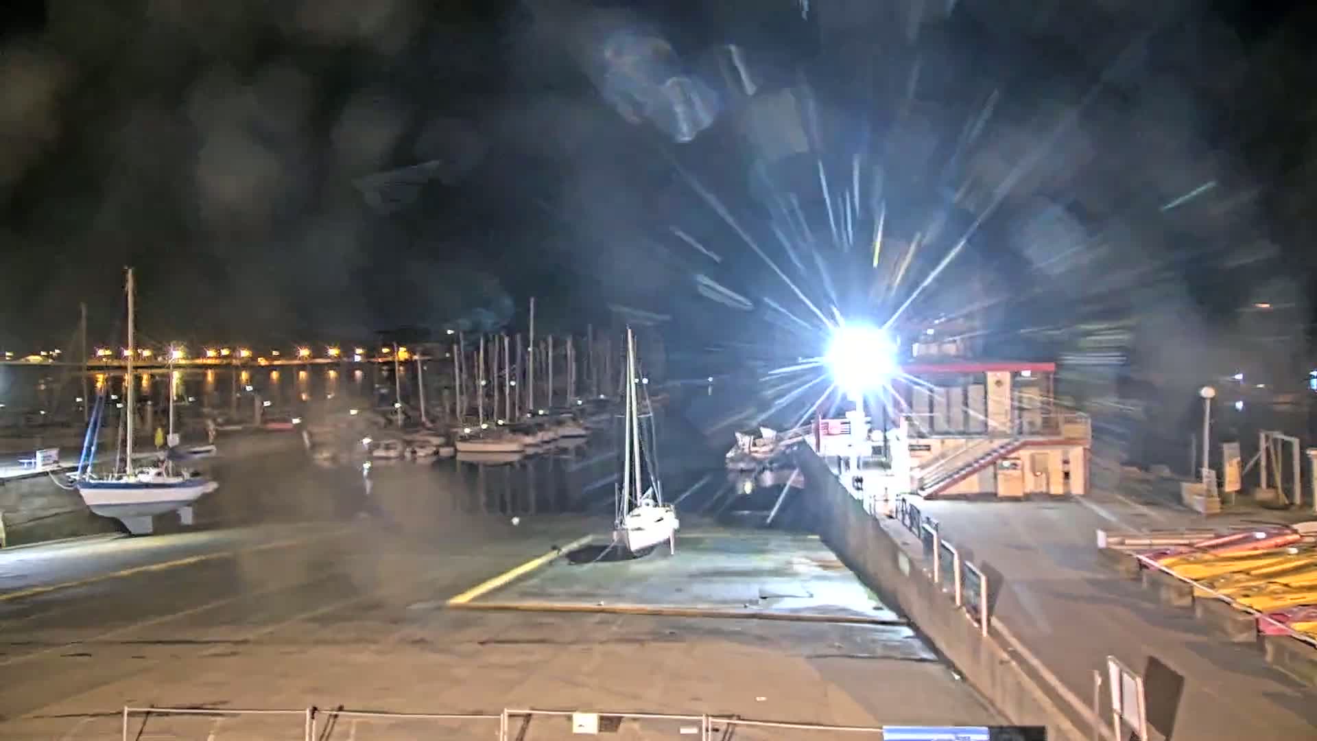 A nighttime view of a marina with numerous sailboats docked, under heavy rain or spray.