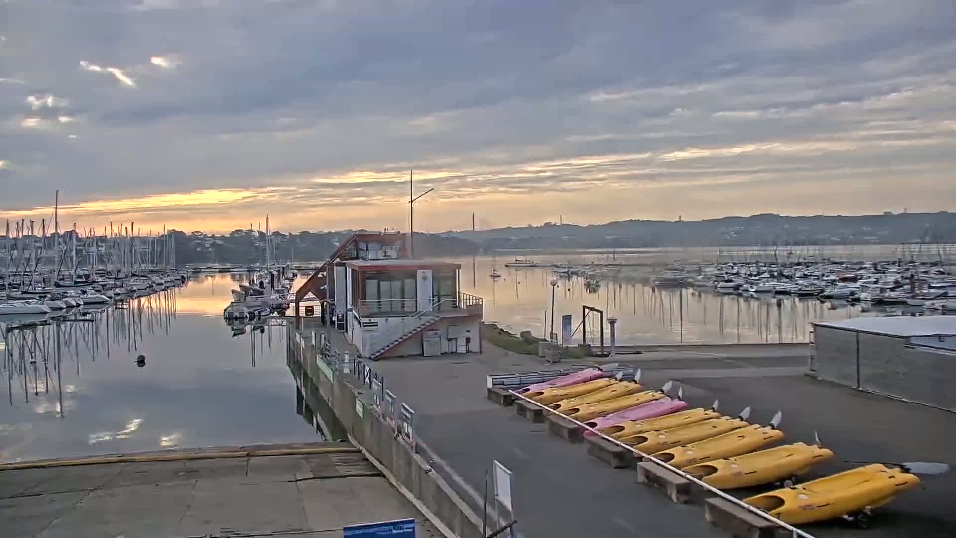 A calm harbor filled with sailboats at sunrise, under a partly cloudy sky, with kayaks lined up on the dock.