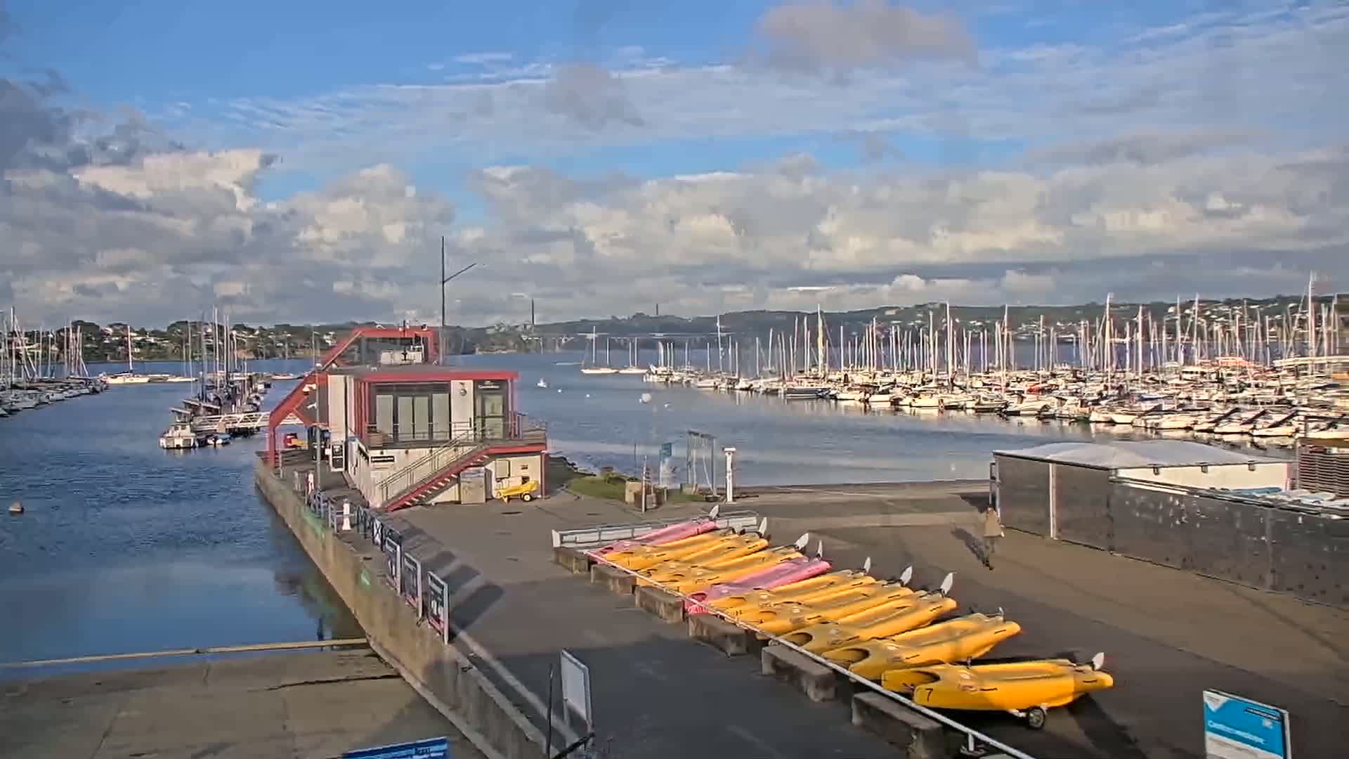 A marina filled with numerous sailboats under a partly cloudy sky, with a red building and rows of yellow and pink kayaks on the shore.