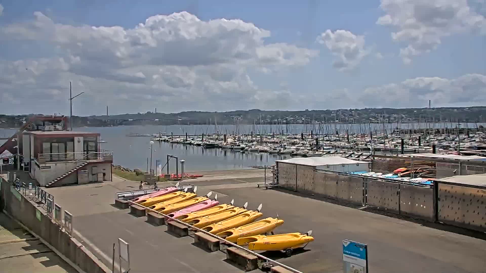 A marina with numerous sailboats and yachts is visible across calm water under a partly cloudy sky, and rows of kayaks are lined up on the shore near a small building.