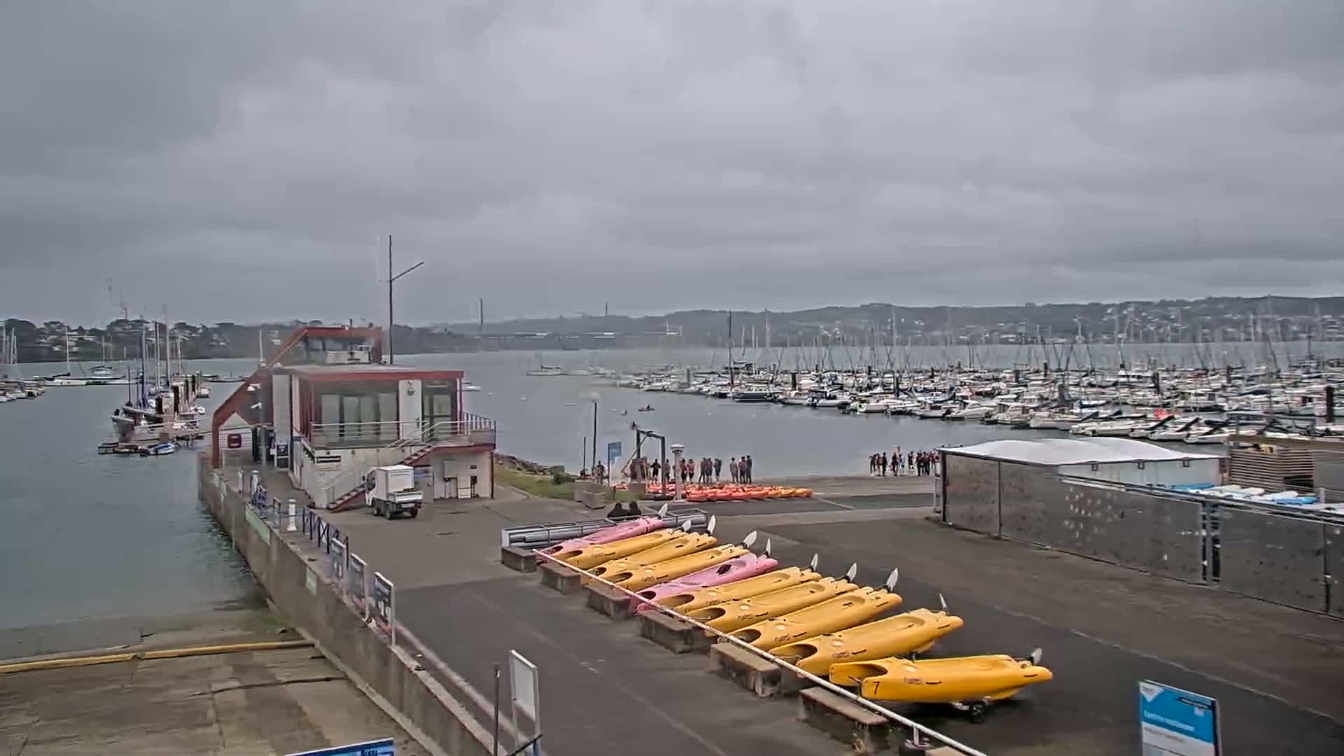 A marina with numerous boats under an overcast sky, a small building, and several kayaks lined up on the shore.