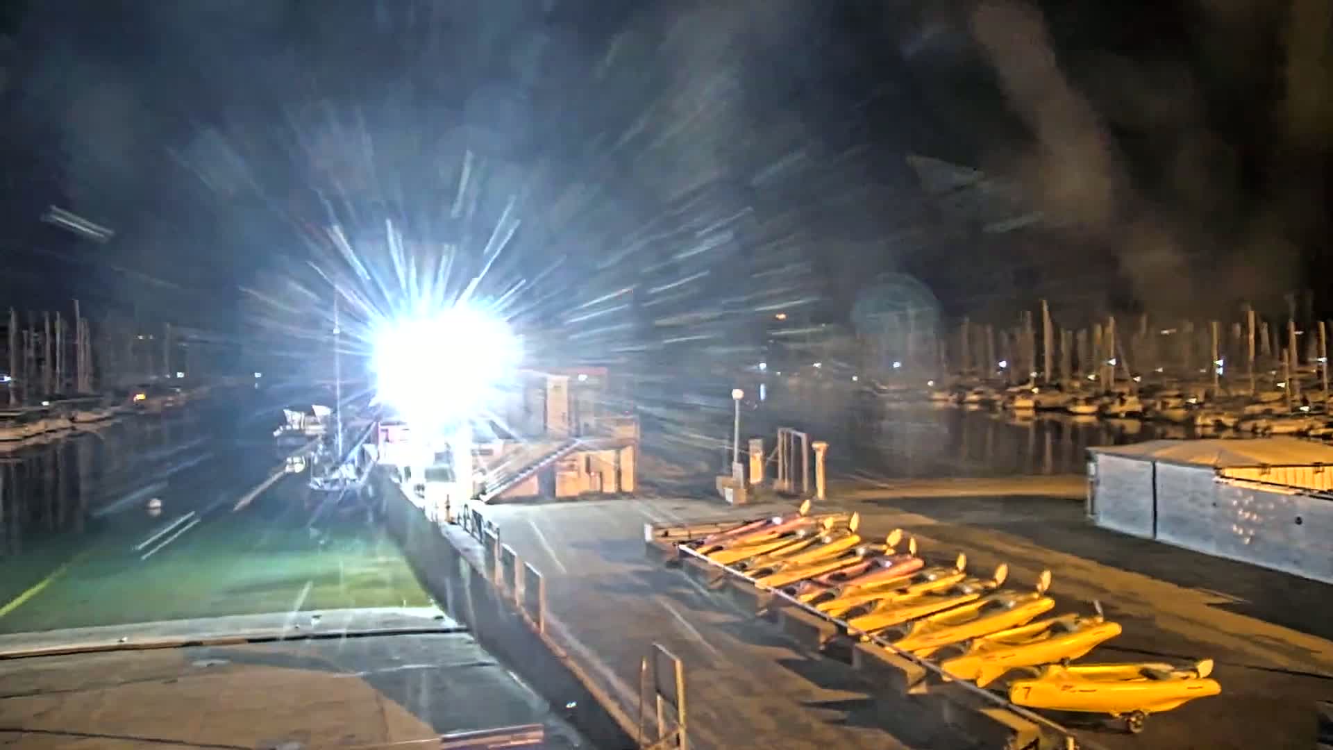 At night, a bright light shines on a dock where several yellow kayaks are lined up, with a marina full of boats visible in the background.
