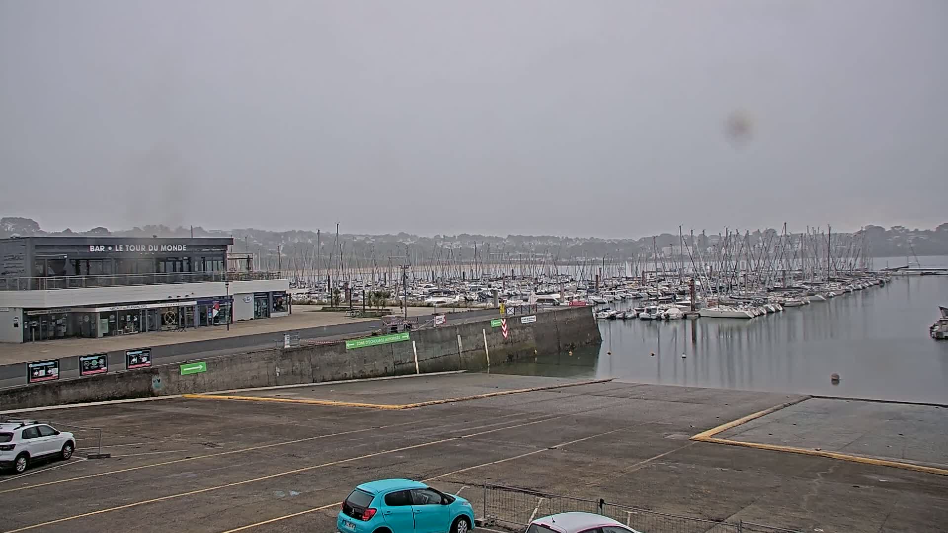 A hazy day reveals a harbor filled with numerous sailboats, a building with a restaurant, and a mostly empty parking lot with a few cars.